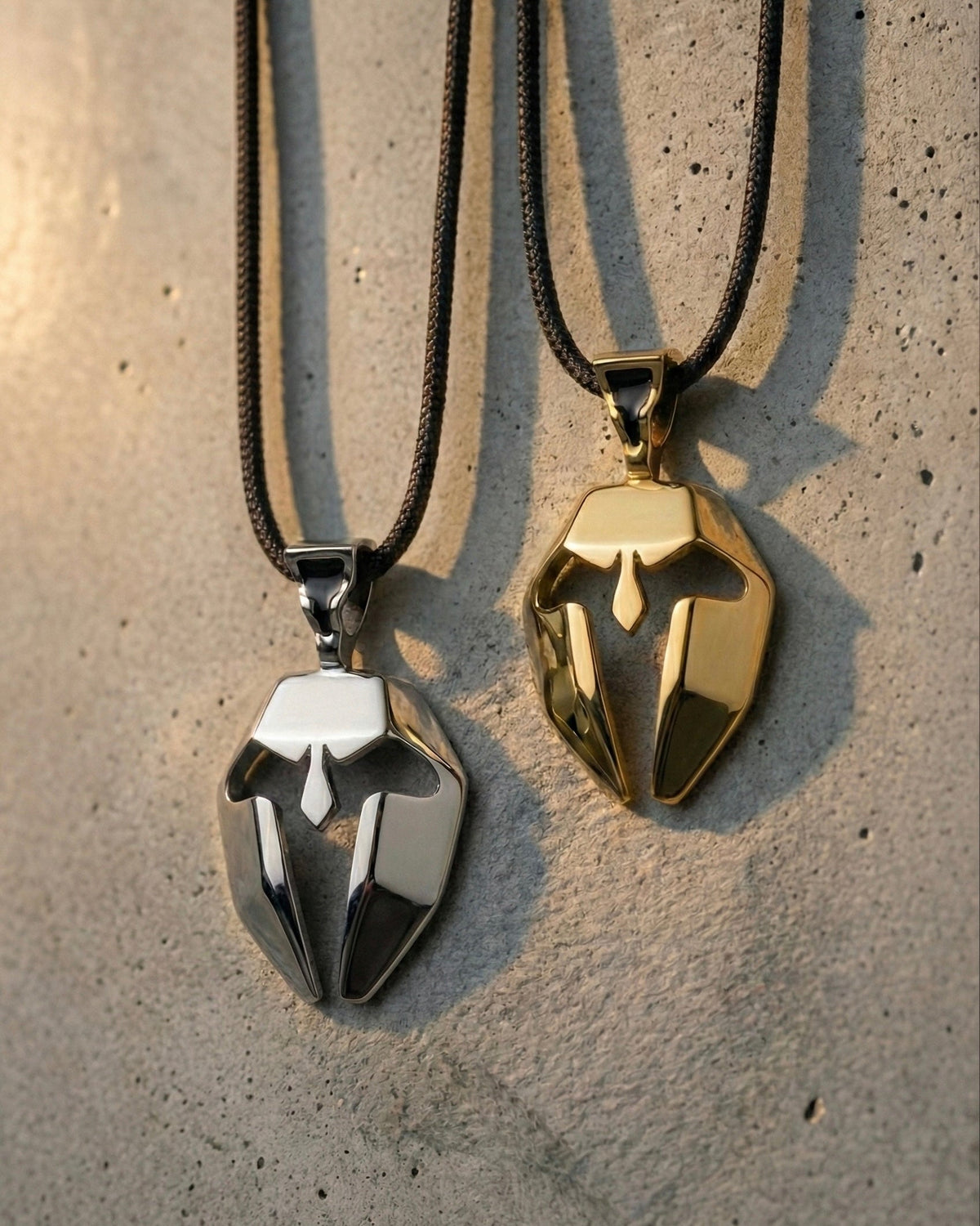 Two metallic Spartan mask pendant necklaces, one gold and one silver, on a textured surface.