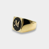 Gold ring with black gemstone and 'M' engraving on a white background