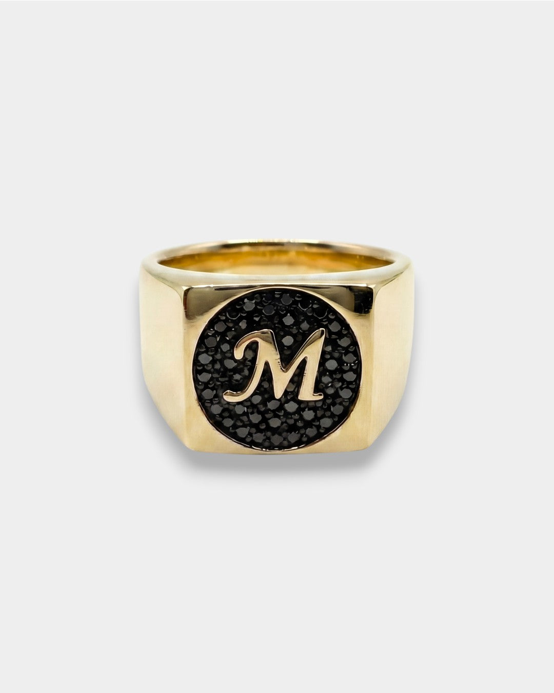 Gold ring with a black circular design and letter 'M' on a white background