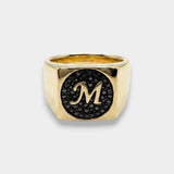 Gold ring with a black circular design and letter 'M' on a white background