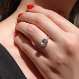 Close-up of a hand wearing a ring with a blue aquamarine gemstone, held against a blurred background.