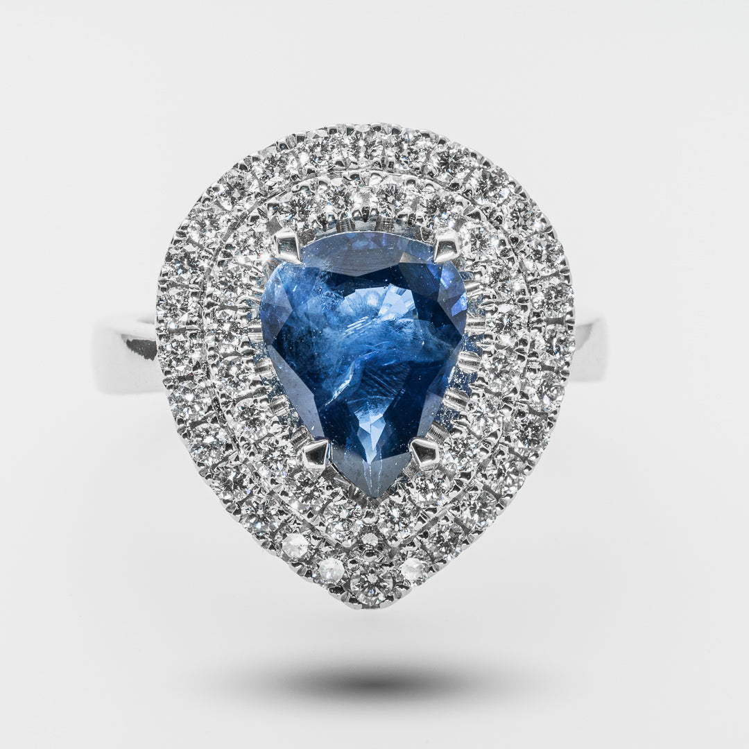 Ring with a pear-shaped blue gemstone and diamond-studded halo on a light gray background