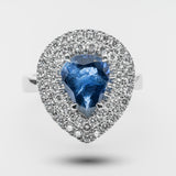 Ring with a pear-shaped blue gemstone and diamond-studded halo on a light gray background