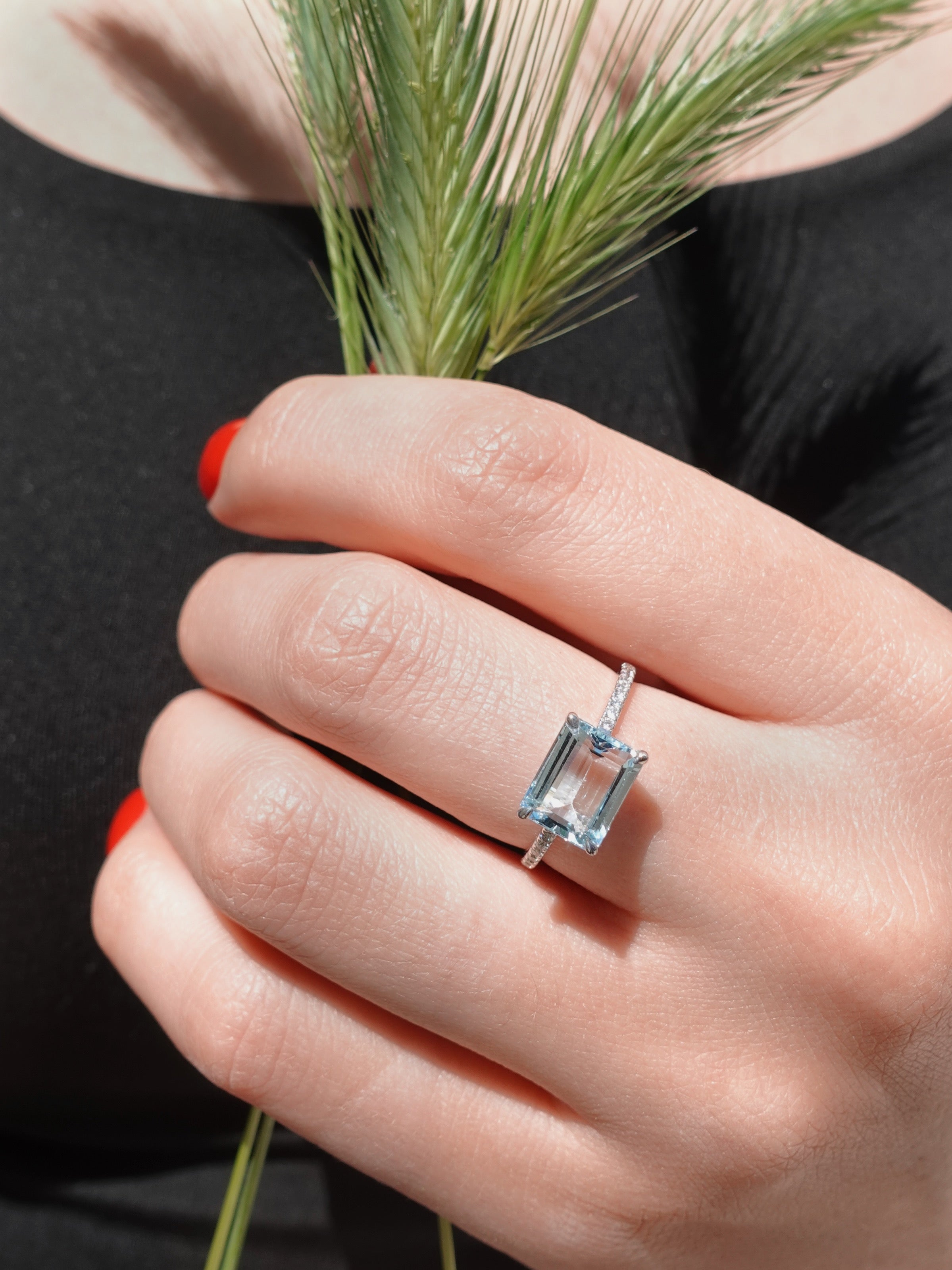 Hand wearing a diamond ring with a blue gemstone, holding a bundle of grass against a black background.