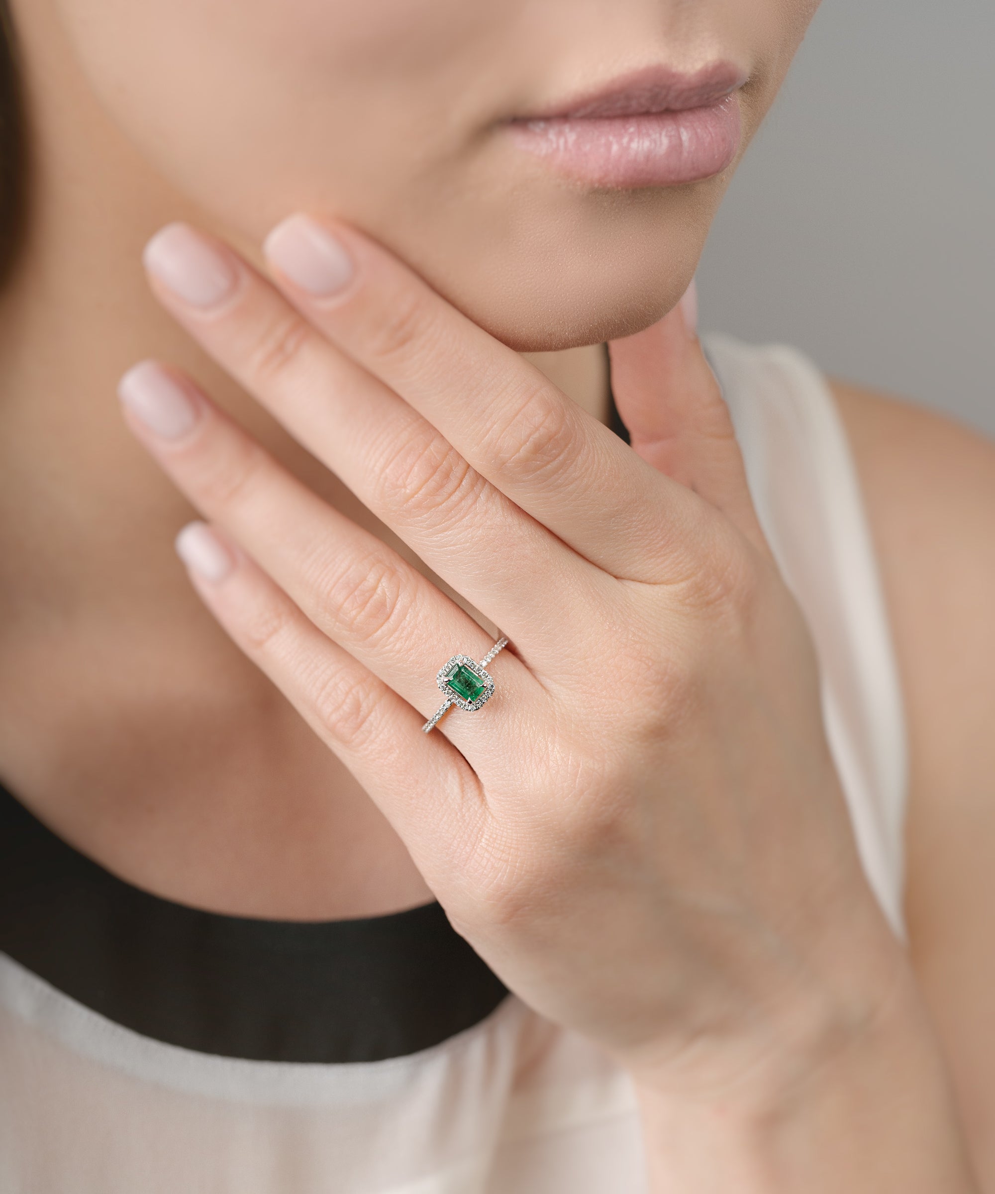 Close-up of a hand wearing a ring with an emerald and diamond halo, held near the face.