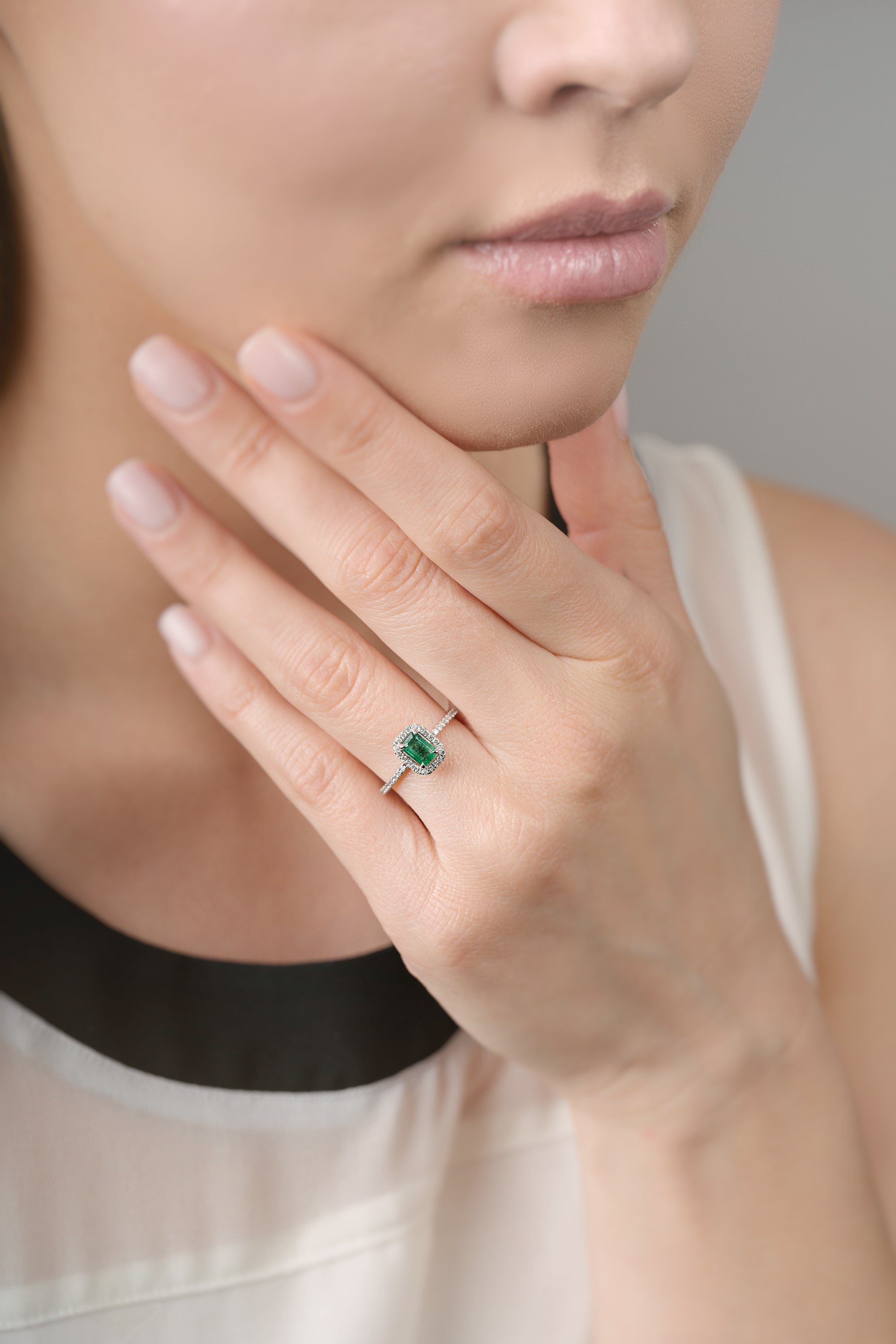 Close-up of a hand wearing a ring with an emerald and diamond halo, held near the face.