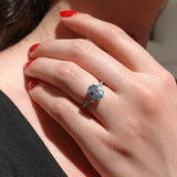 Close-up of a hand wearing a ring with a blue gemstone, against a blurred background.