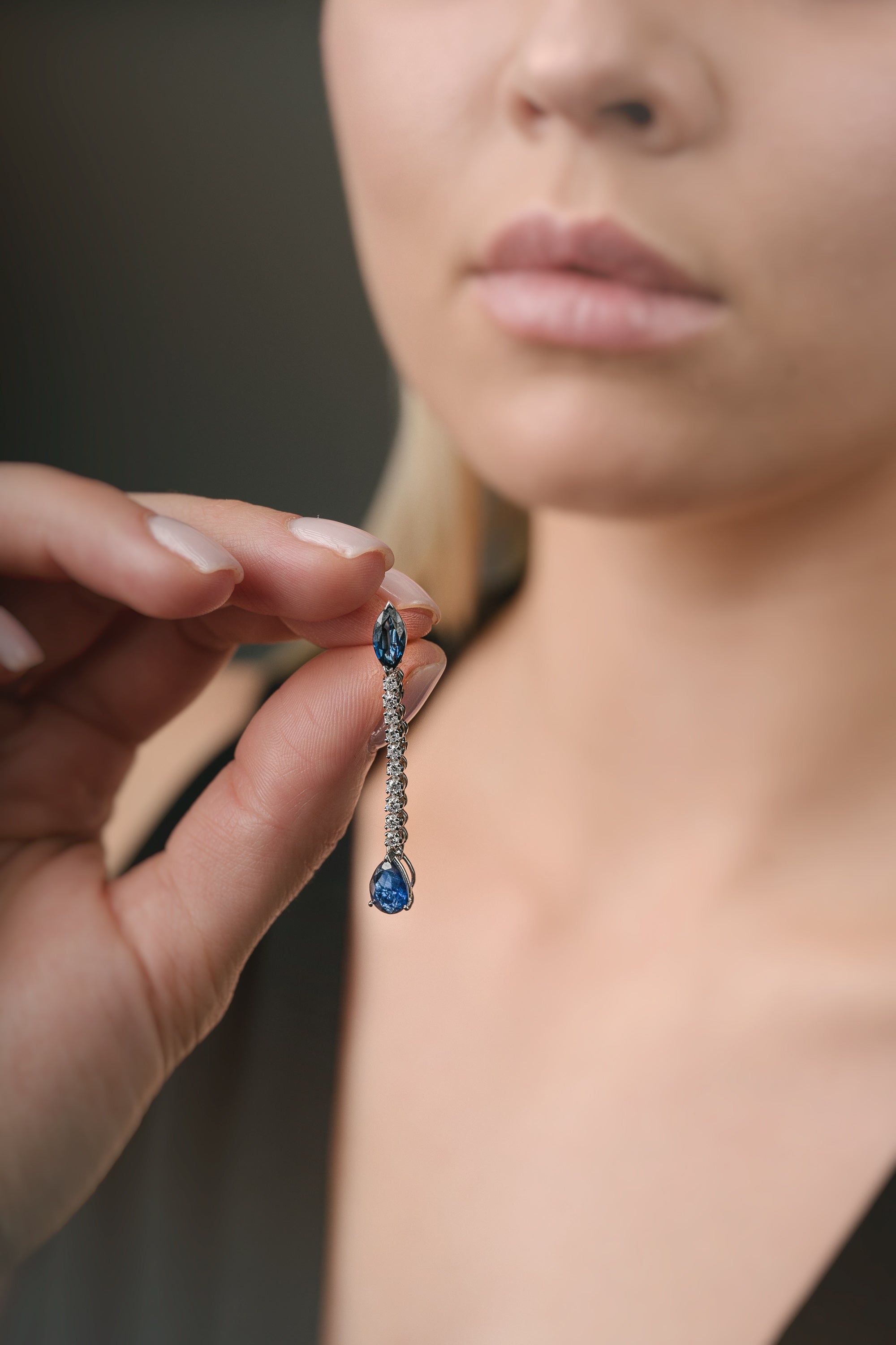 Close-up of a hand holding a blue sapphire gemstone earring near a person's face.