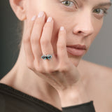 A woman wearing a ring with green and blue gemstones, touching her face.
