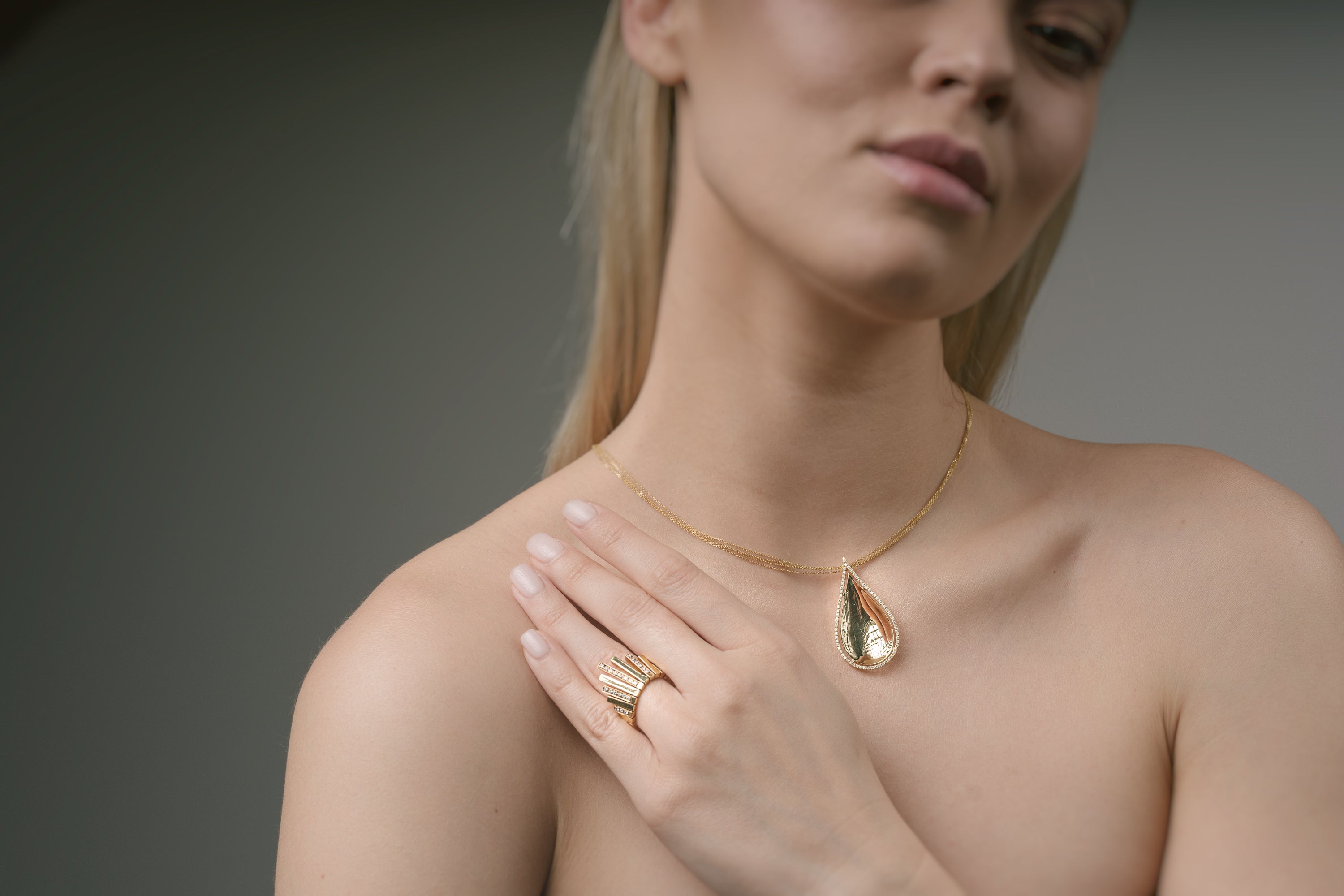 Woman wearing a gold teardrop necklace and sunburst ring on a neutral background