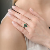 Close-up of a hand wearing a ring with a pink gemstone and emeralds, touching the shoulder against a neutral background