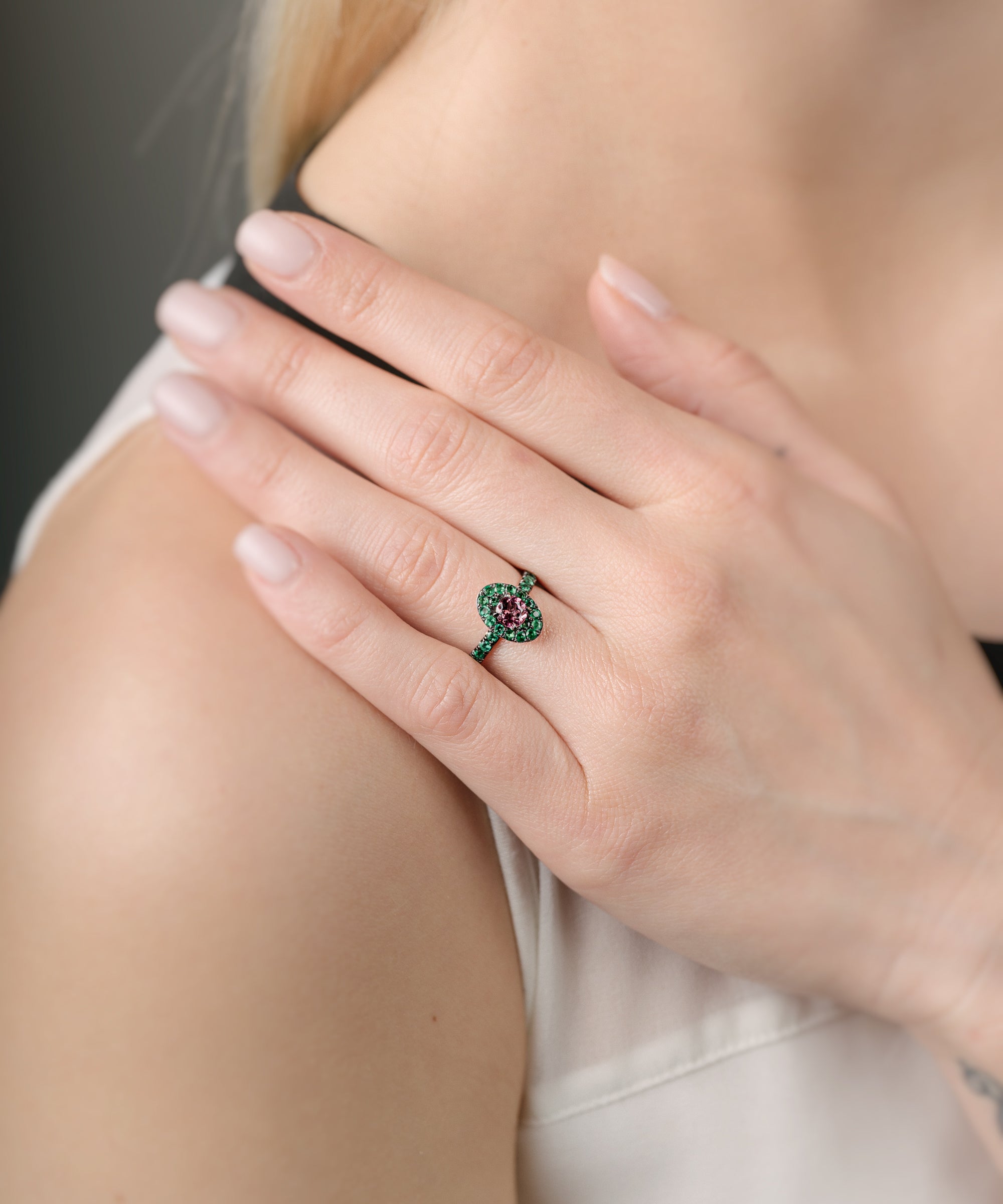 Close-up of a hand wearing a ring with a pink gemstone and emeralds, touching the shoulder against a neutral background