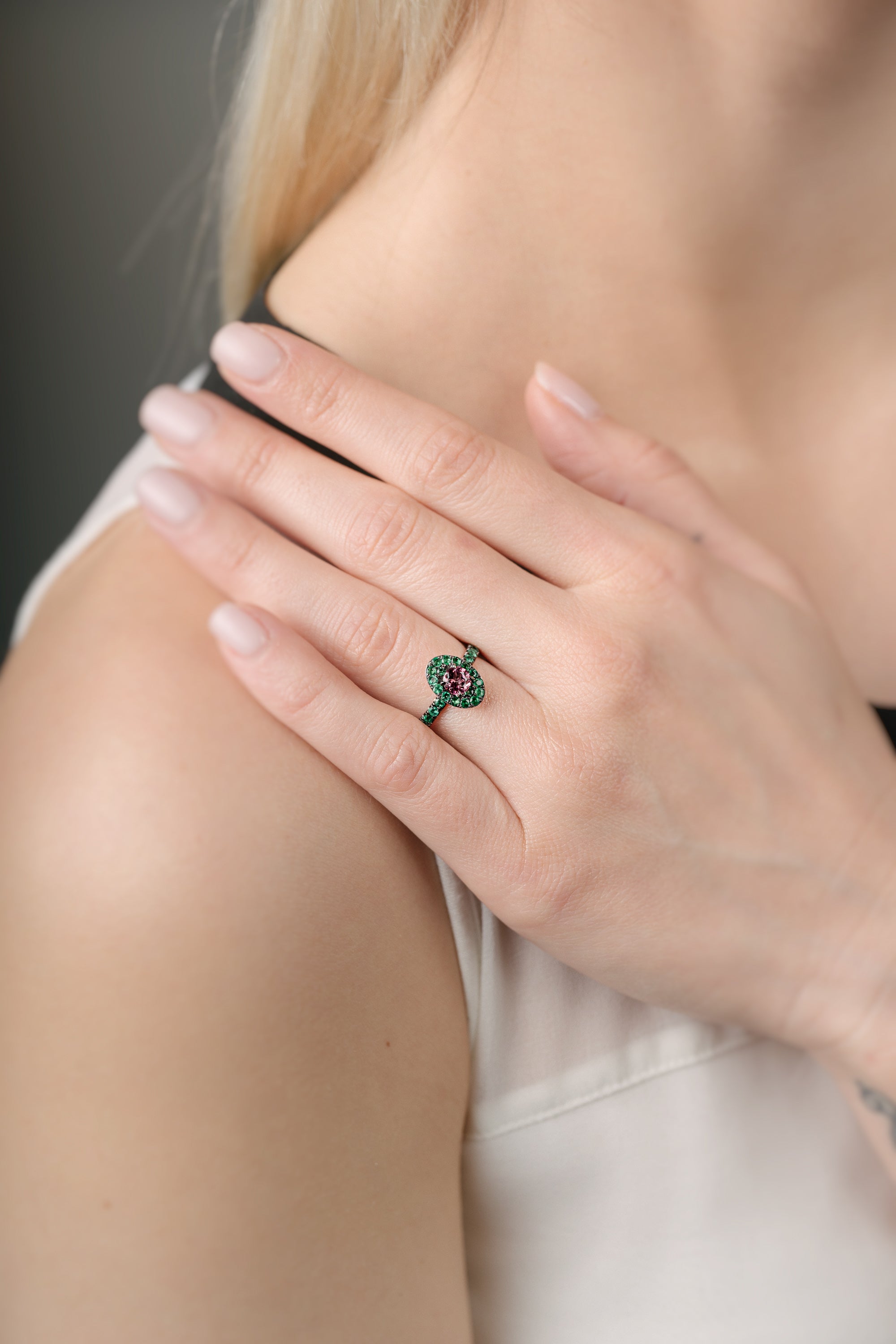 Close-up of a hand wearing a ring with a pink gemstone and emeralds, touching the shoulder against a neutral background
