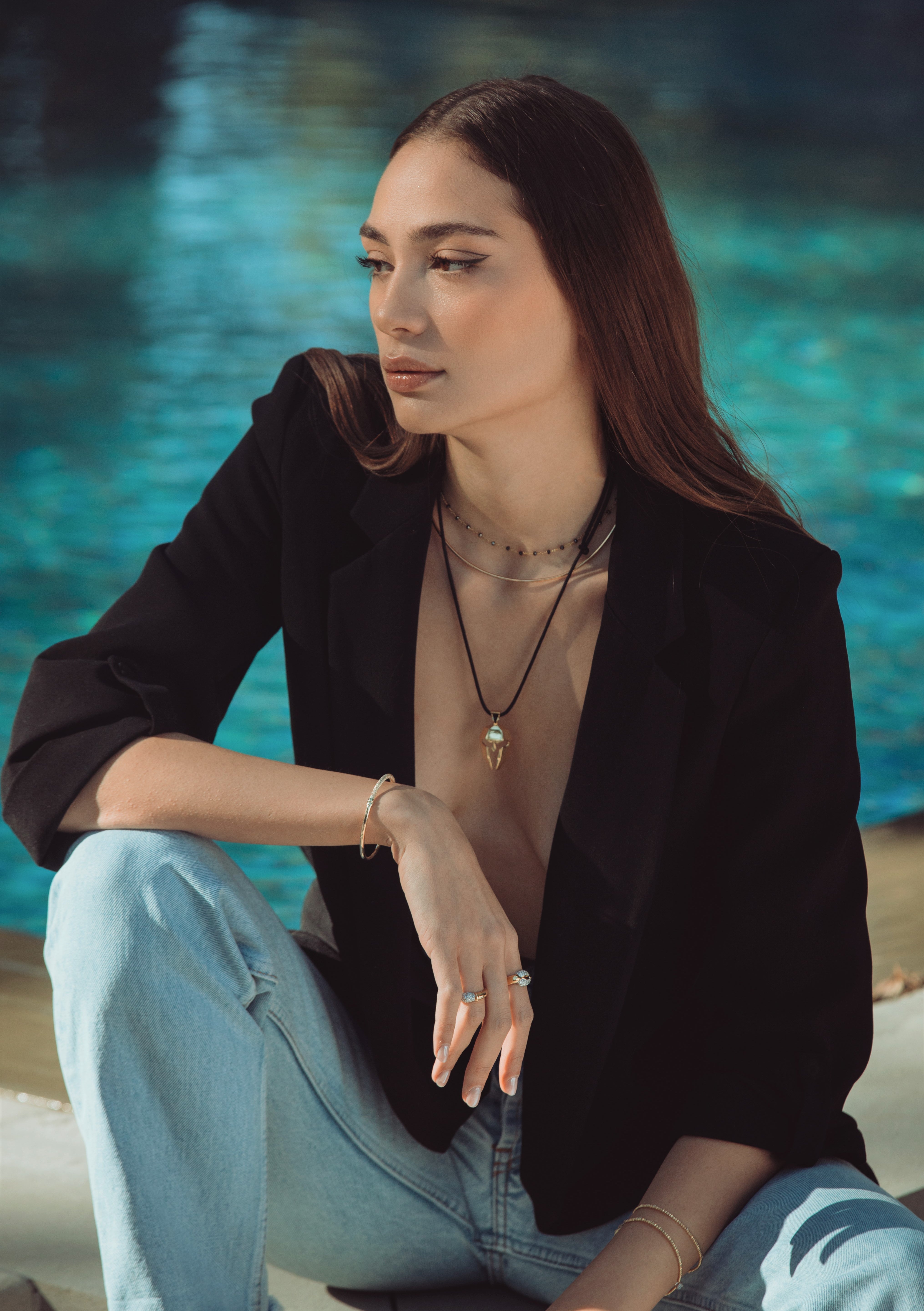 Woman sitting by a pool wearing a black blazer and light blue jeans.