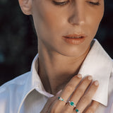 Woman wearing a white shirt with a close-up of her hand holding a ring with green emerald gemstones.