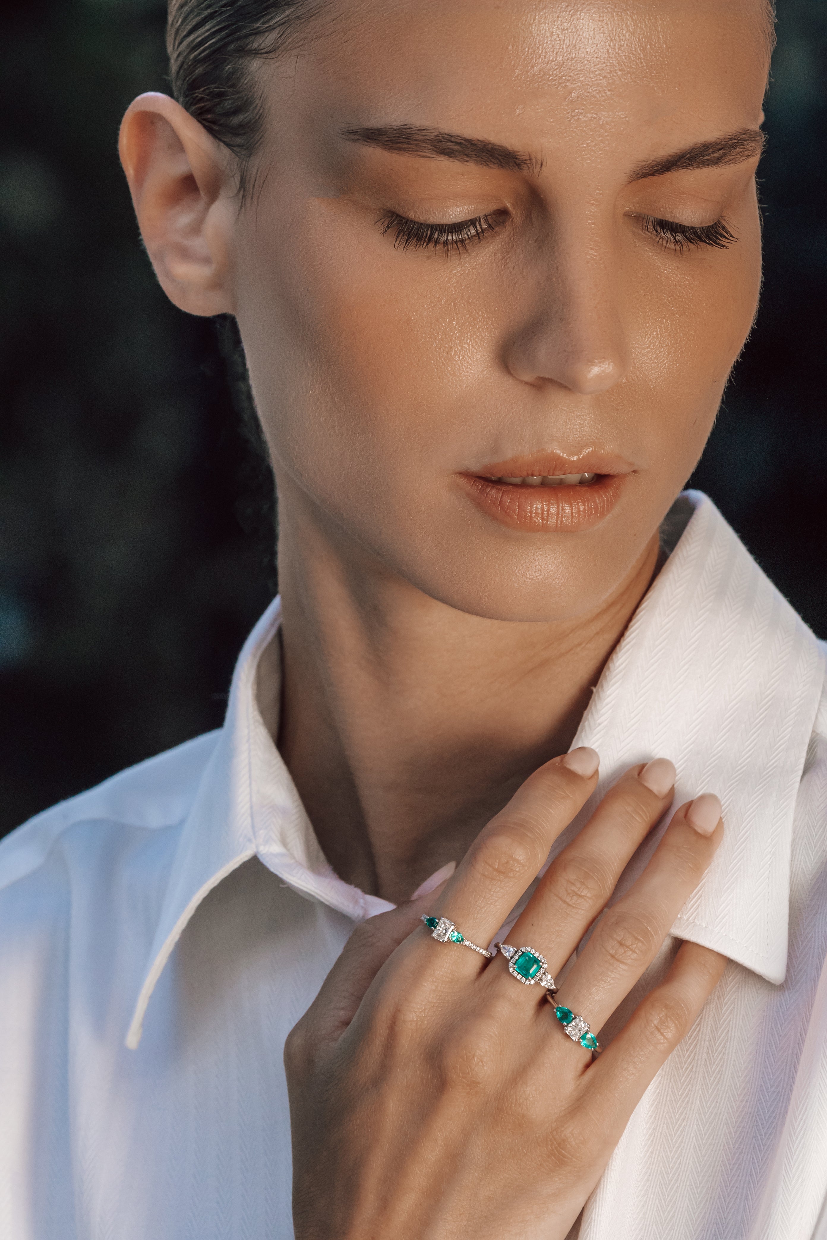 Woman wearing a white shirt with a close-up of her hand holding a ring with green emerald gemstones.