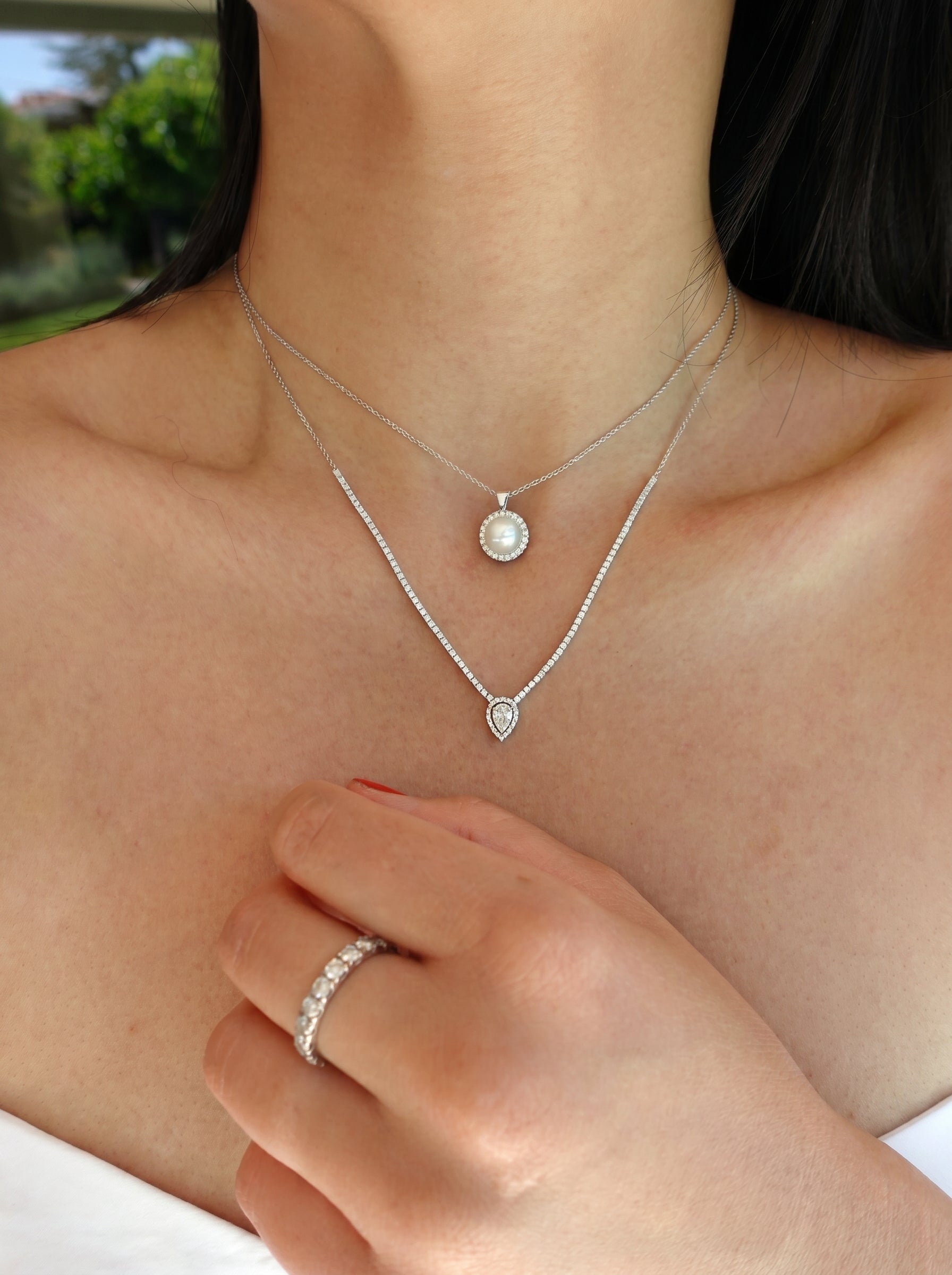 Close-up of a person wearing two white-gold pearl necklaces with a blurred outdoor background