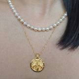 Gold coin necklace with an angel image worn by a person, with a pearl necklace layered on top.