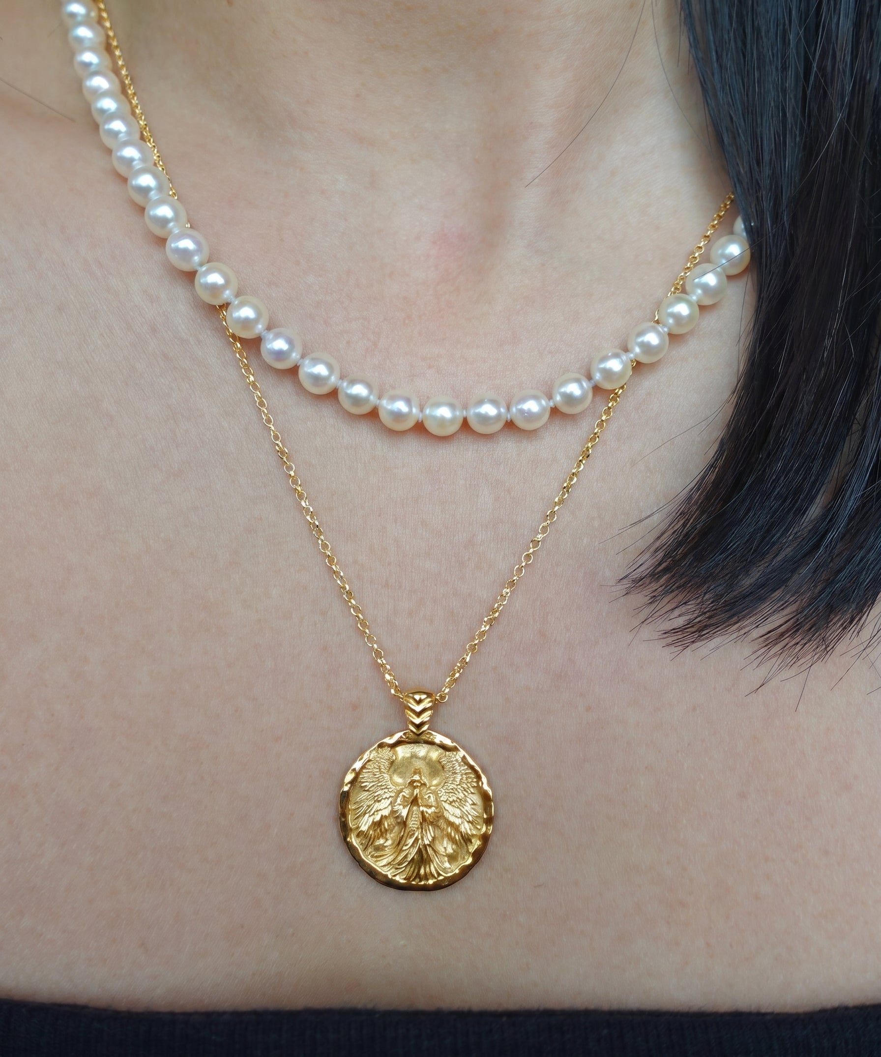 Gold coin necklace with an angel image worn by a person, with a pearl necklace layered on top.
