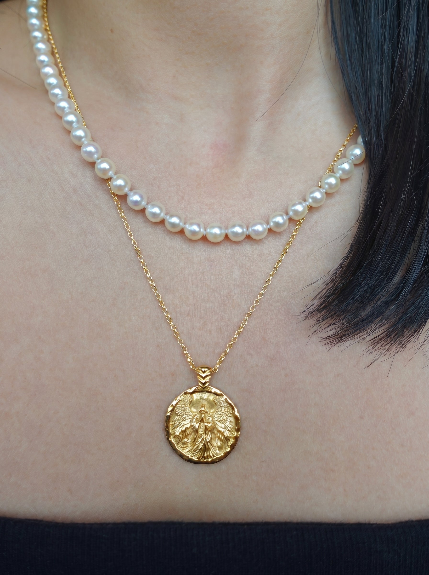 Gold coin necklace with an angel image worn by a person, with a pearl necklace layered on top.