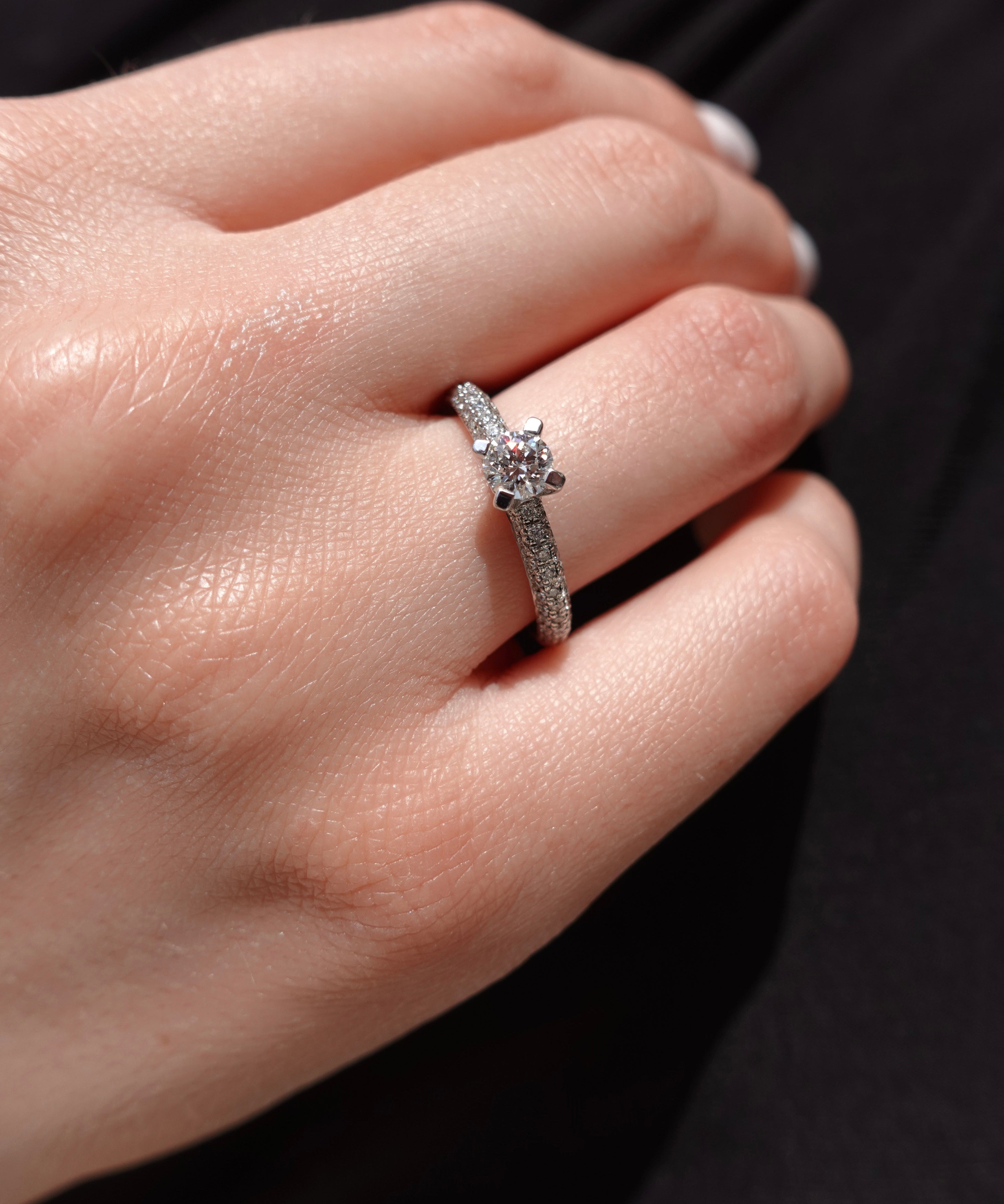 a hand wearing a brilliant-cut diamond engagement ring with pave diamonds on the shank. 