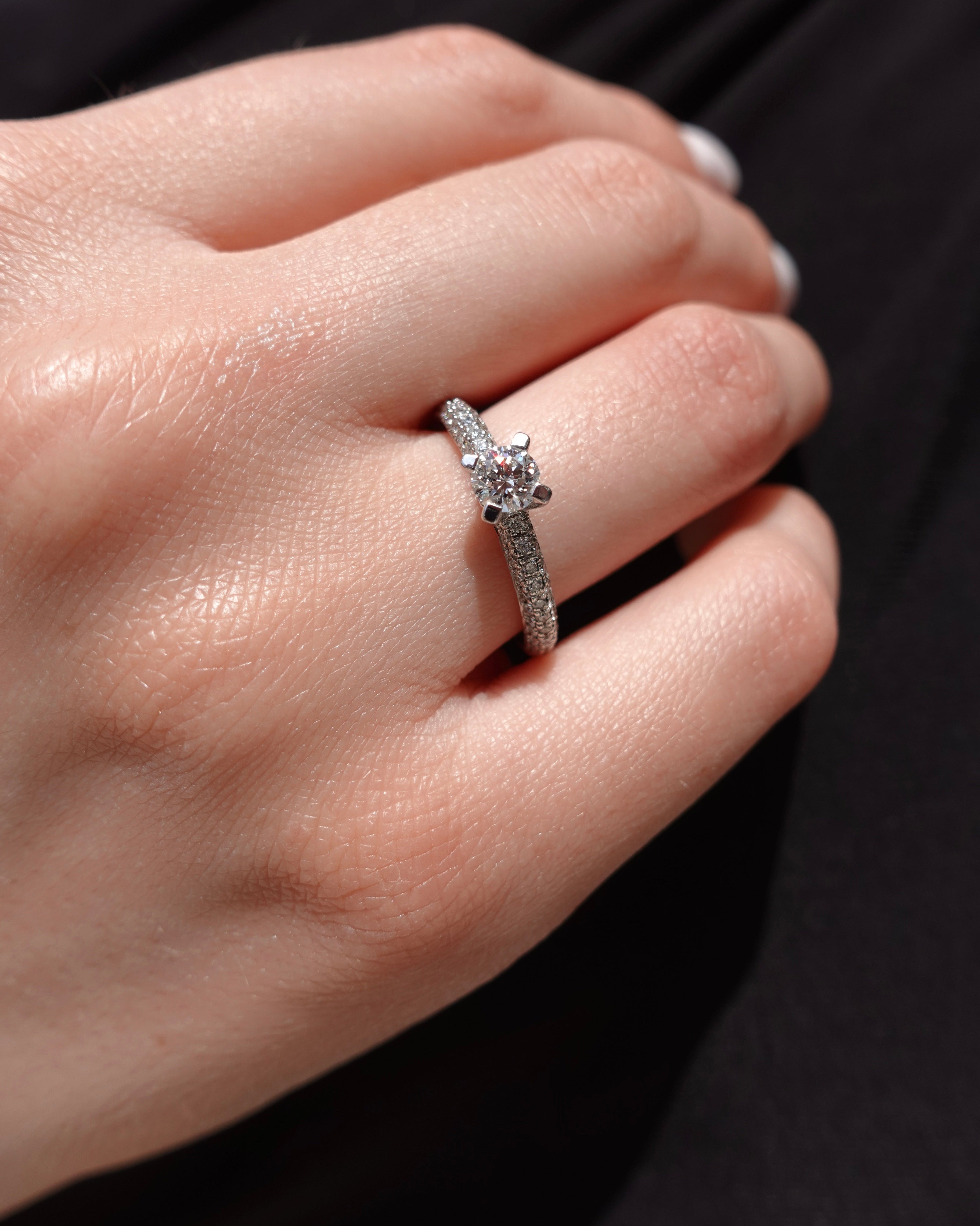 a hand wearing a brilliant-cut diamond engagement ring with pave diamonds on the shank. 
