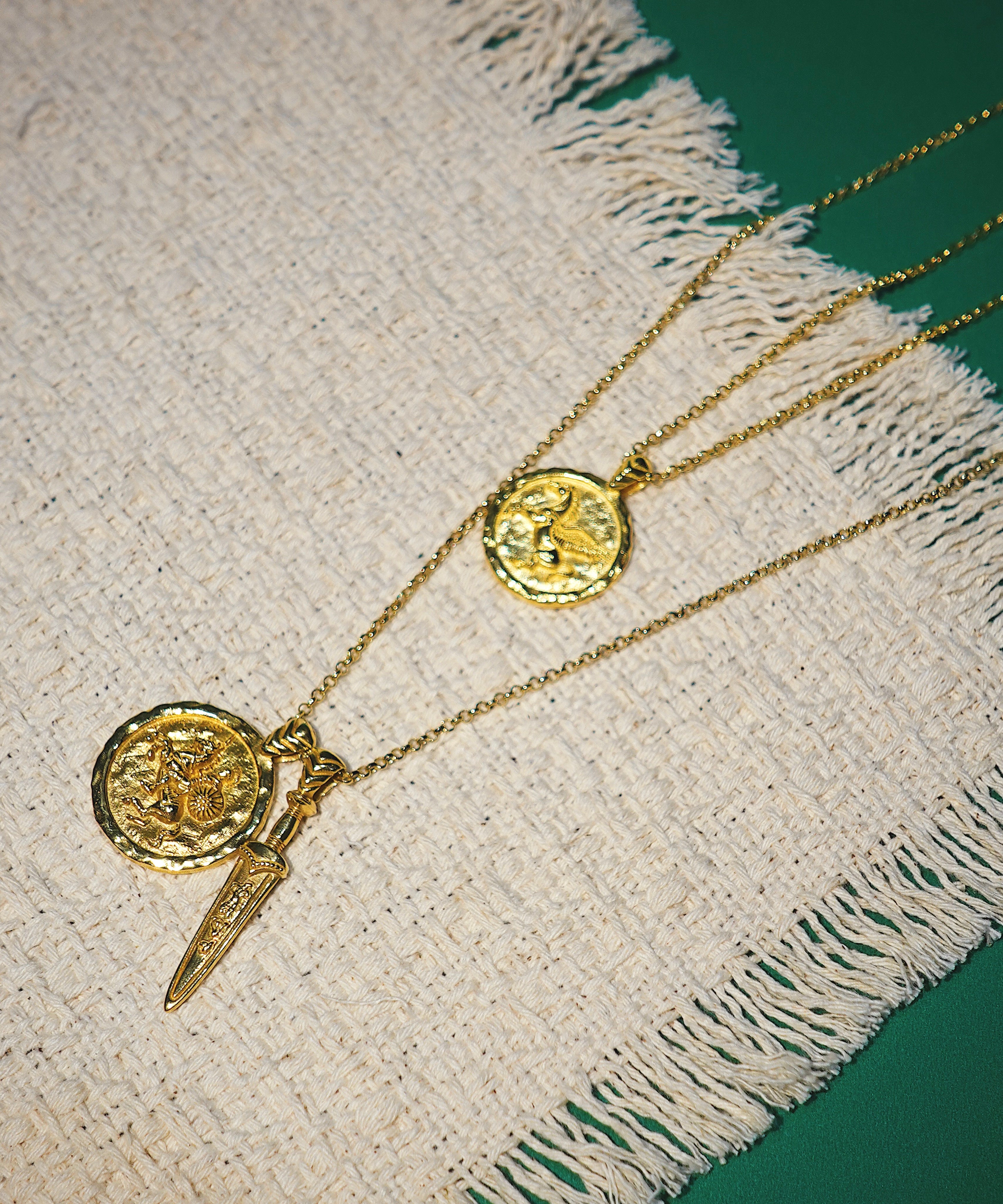 Two gold necklaces with coin pendants on a textured fabric surface.