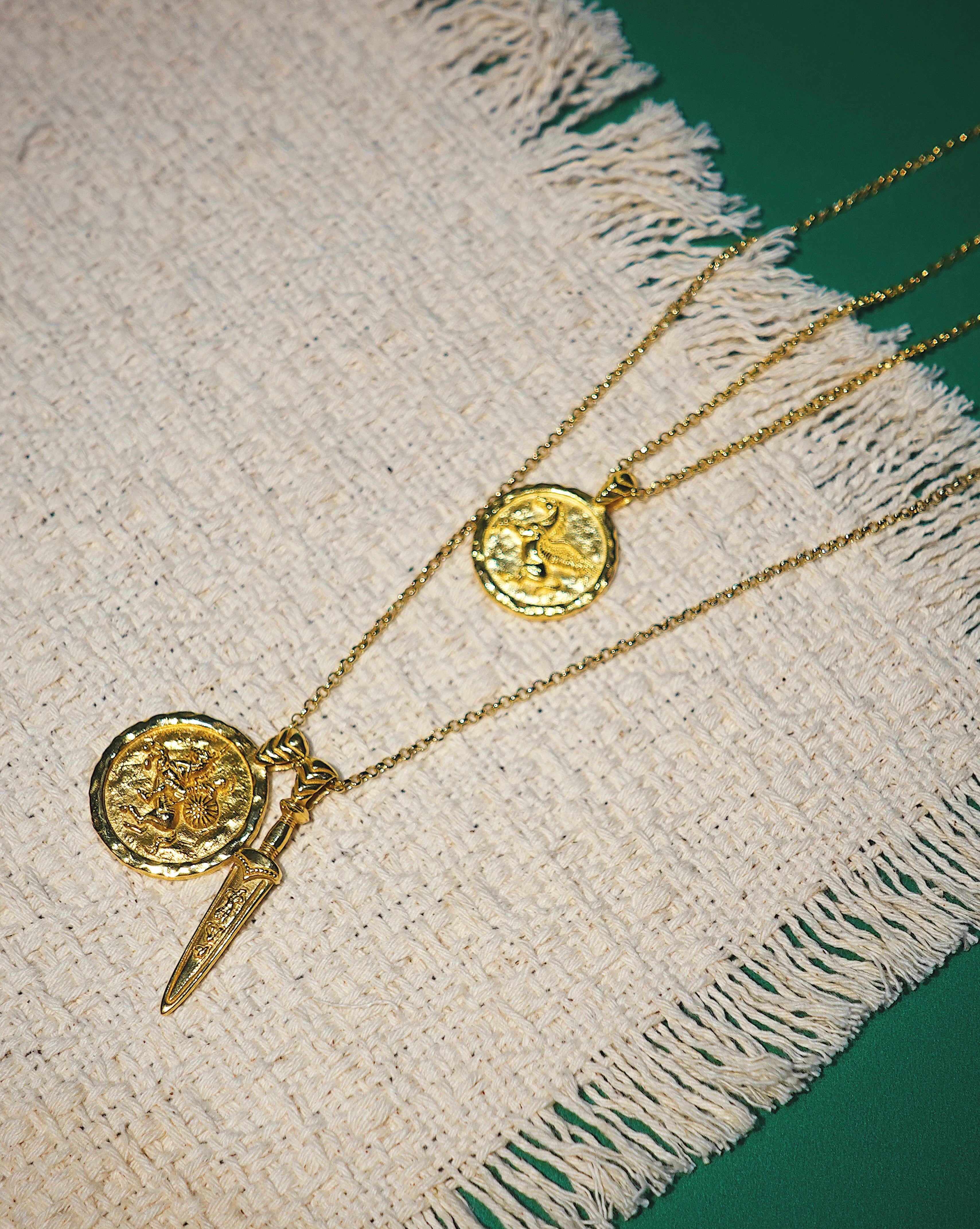 Two gold necklaces with coin pendants on a textured fabric surface.