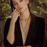 Woman wearing a black blazer with layered necklaces against a blurred natural background