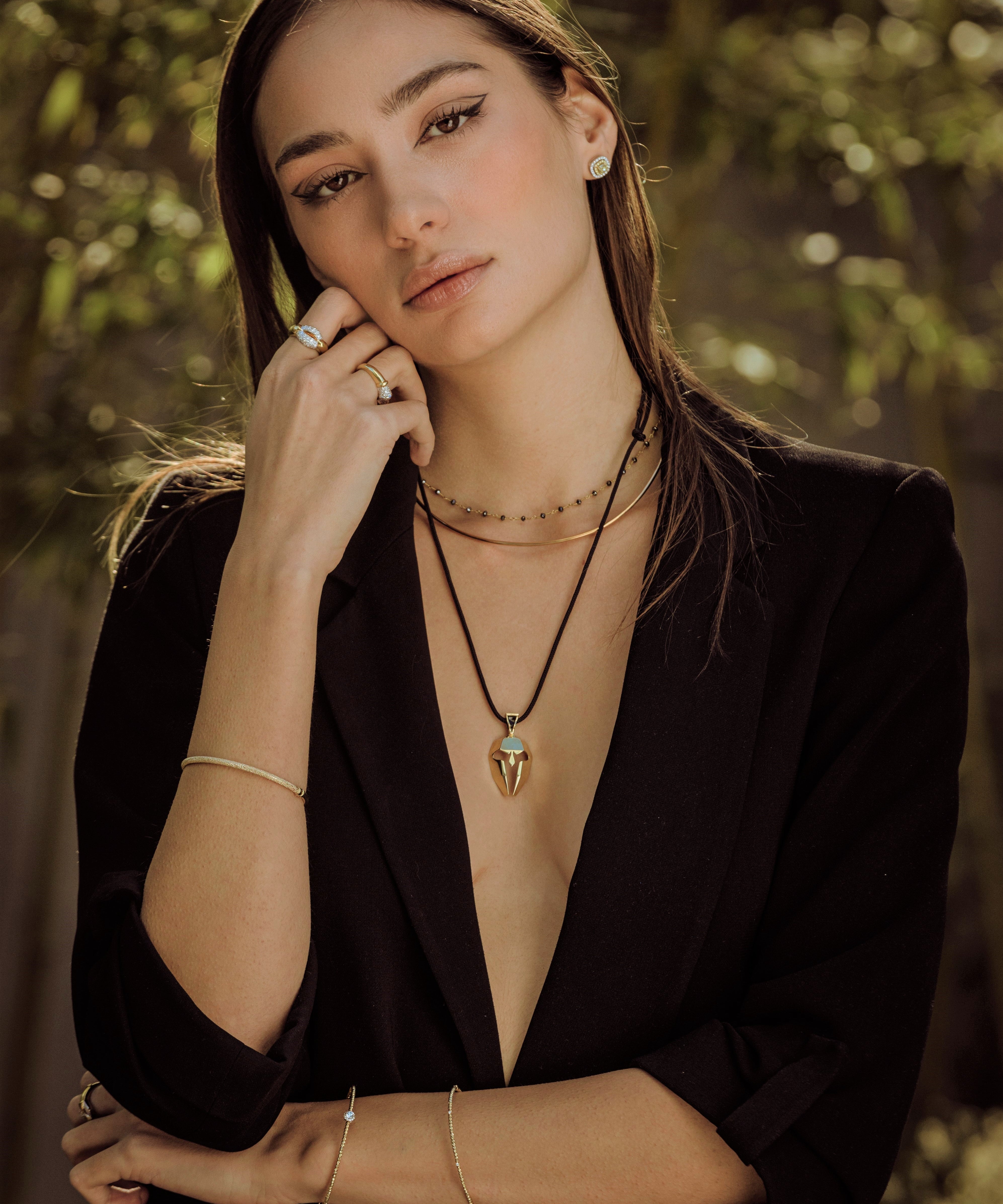 Woman wearing a black blazer with layered necklaces against a blurred natural background