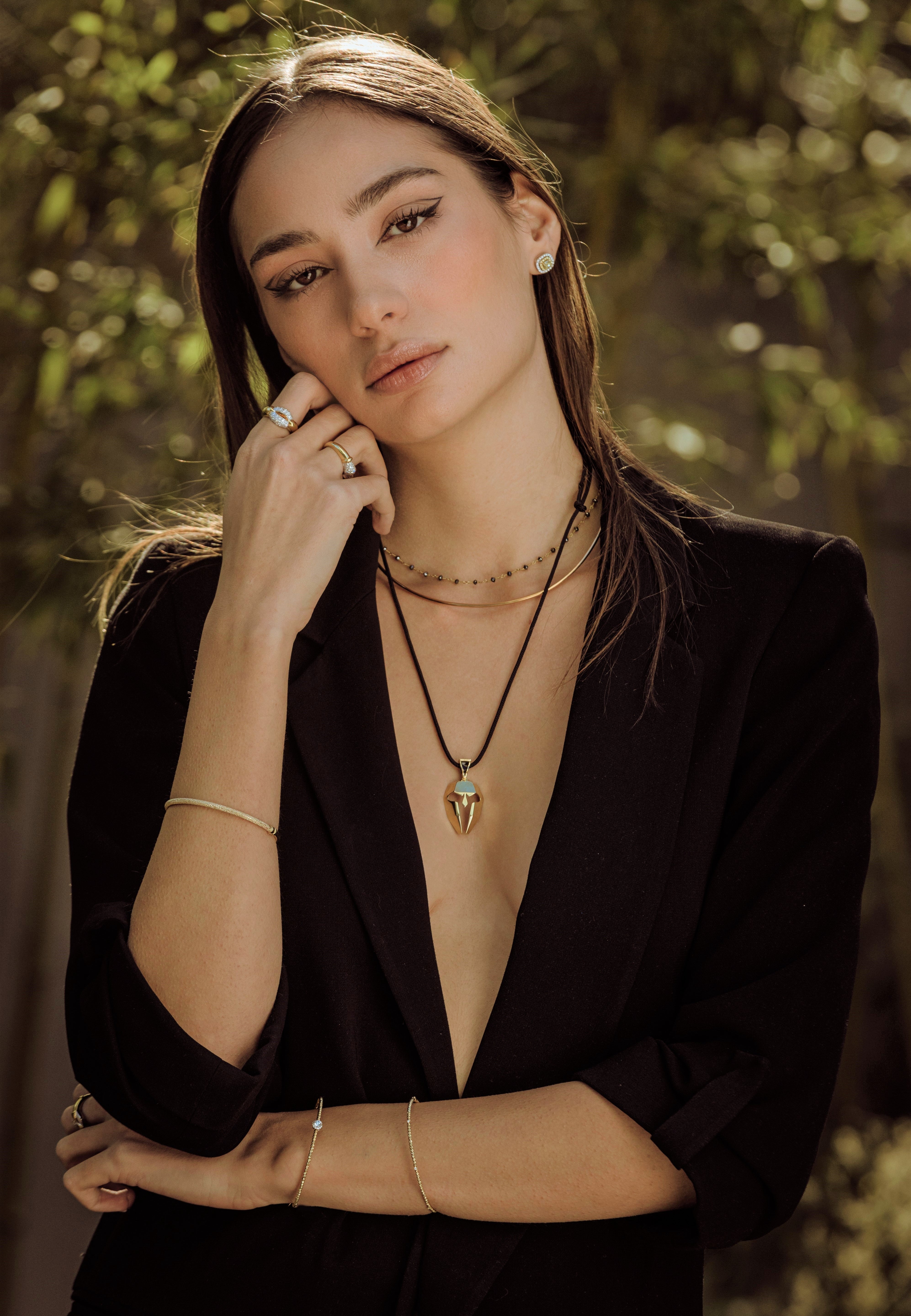 Woman wearing a black blazer with layered necklaces against a blurred natural background