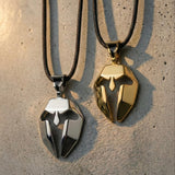 Two metallic Spartan mask pendant necklaces, one gold and one silver, on a textured surface.