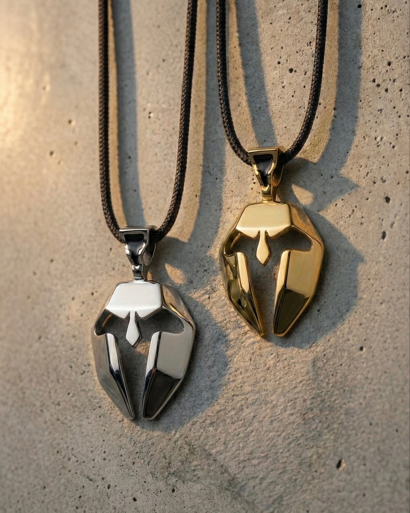 Two metallic Spartan mask pendant necklaces, one gold and one silver, on a textured surface.