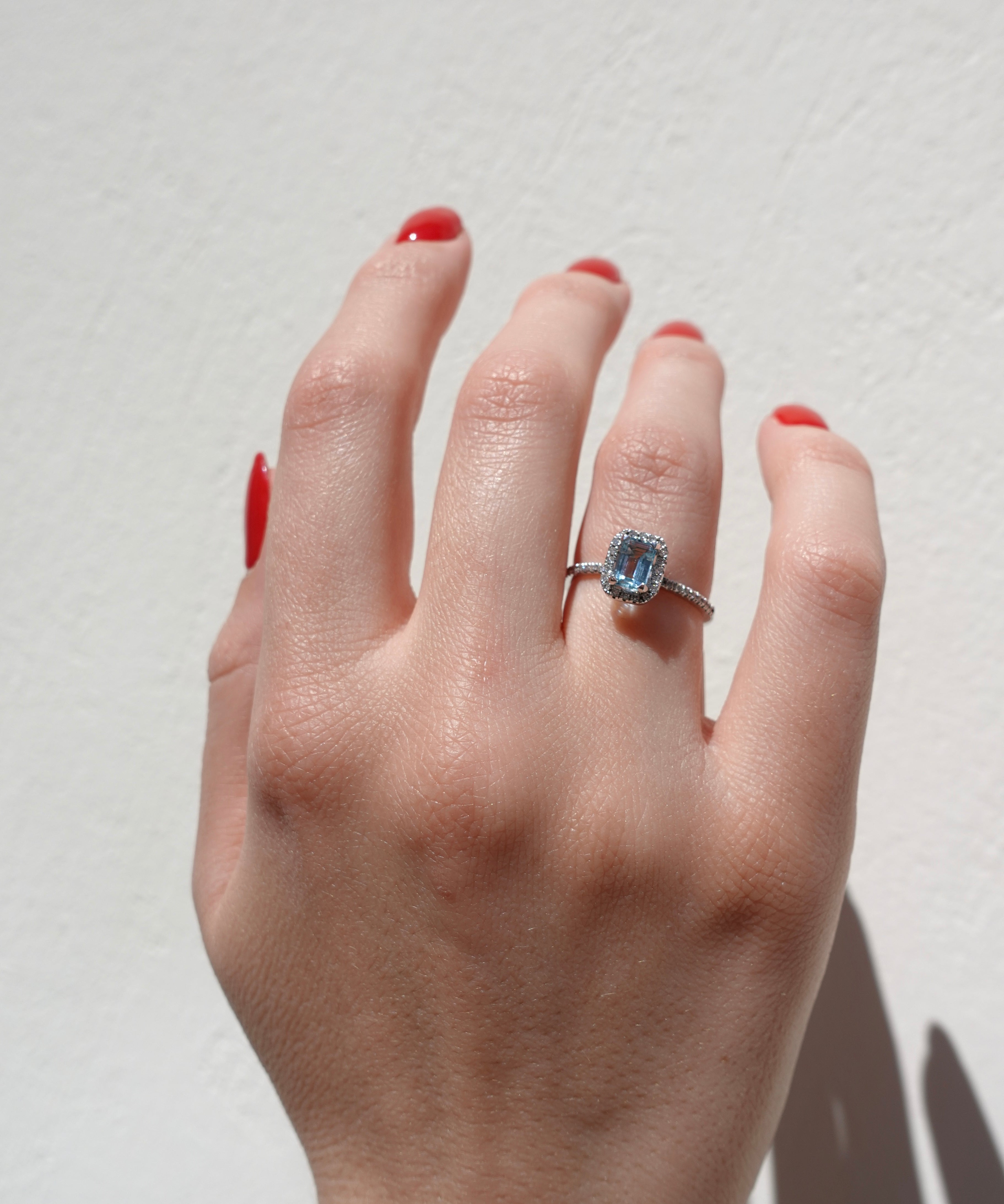 Hand wearing a diamond halo ring with a center aquamarine gemstone on a plain background