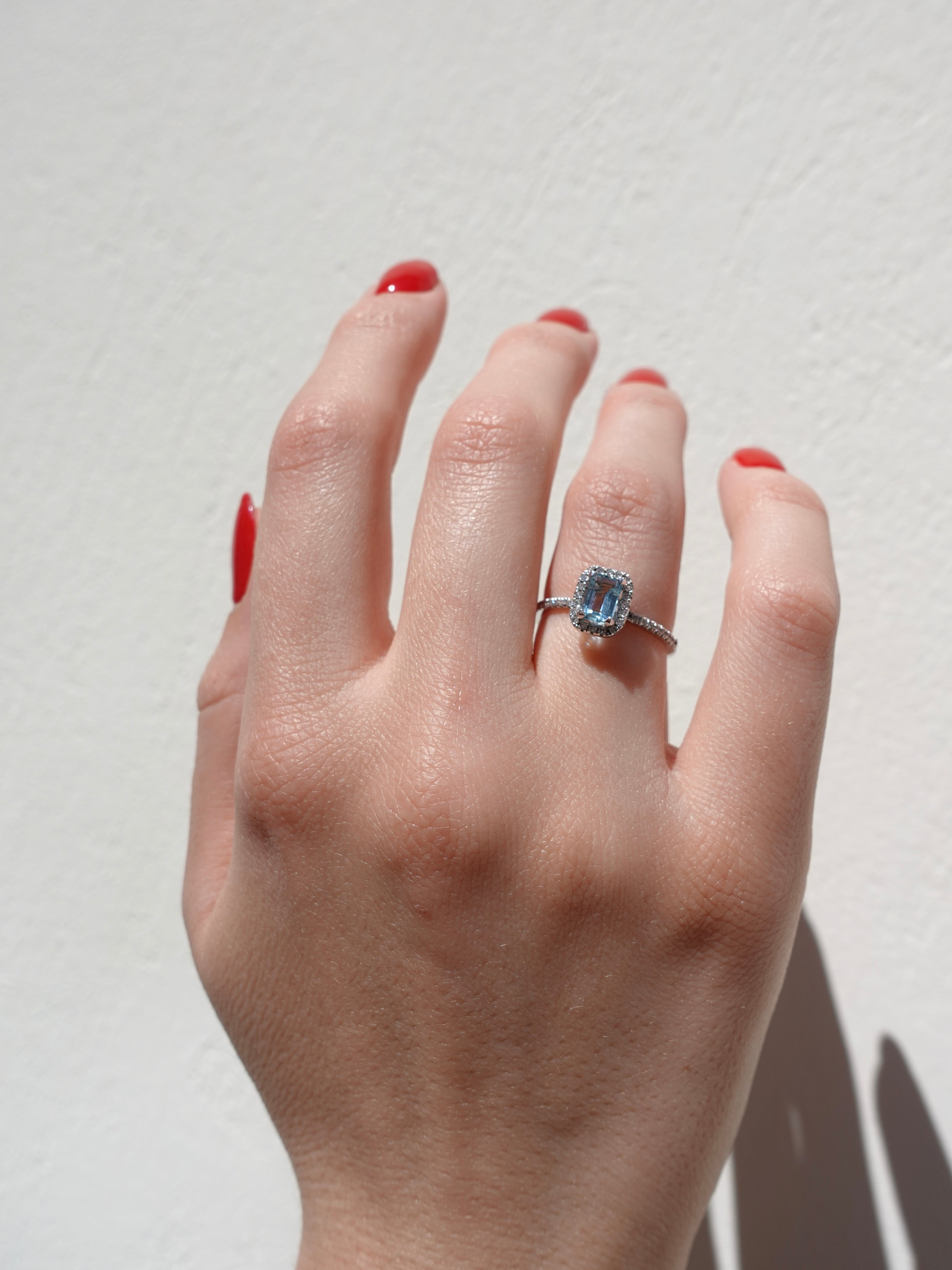 Hand wearing a diamond halo ring with a center aquamarine gemstone on a plain background