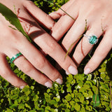 Two hands with green gemstone rings on a green leafy background