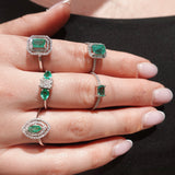 Hand wearing multiple emerald and diamond rings on a black background