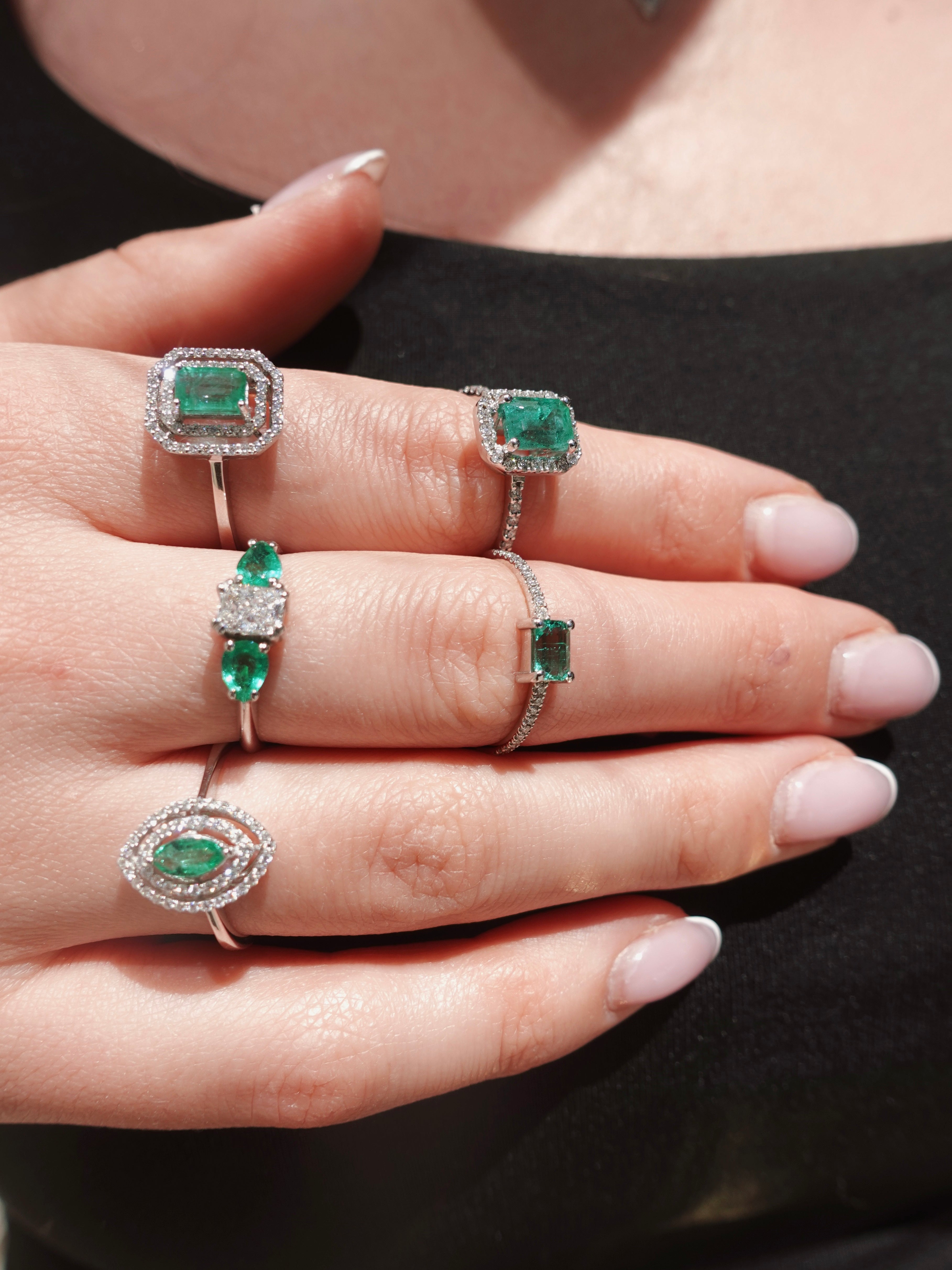 Hand wearing multiple emerald and diamond rings on a black background