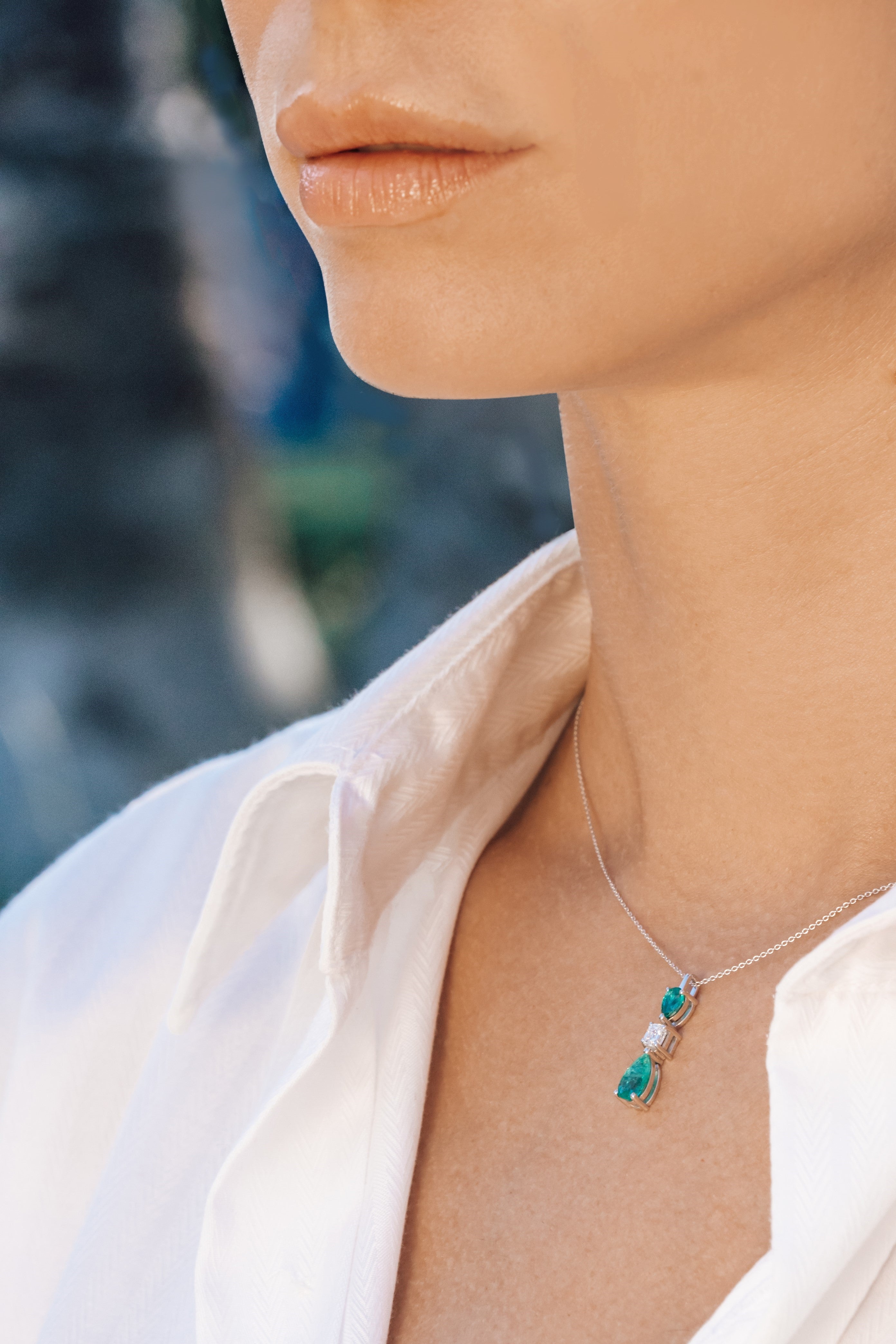 Necklace with an emerald and diamond pendant worn by a person against a blurred outdoor background