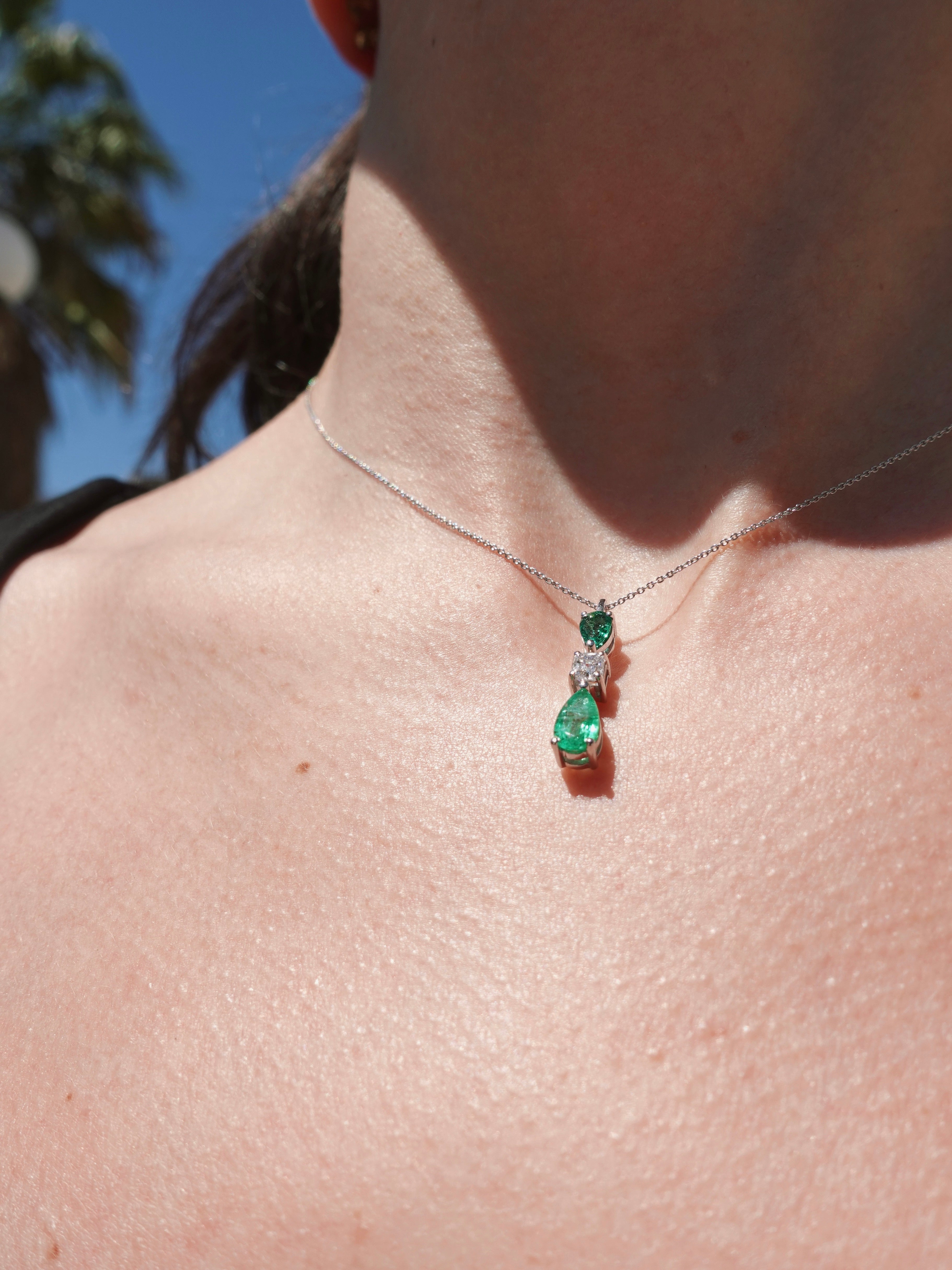 Close-up of a necklace with a green pendant on a person's neck against a blurred outdoor background.