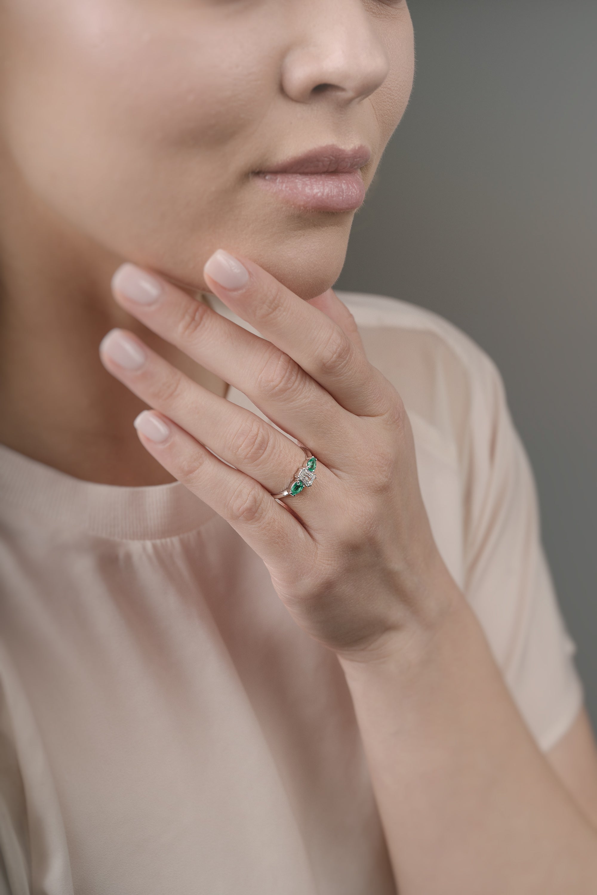 Close-up of a woman's hand wearing a ring with emeralds and diamond, touching her face.