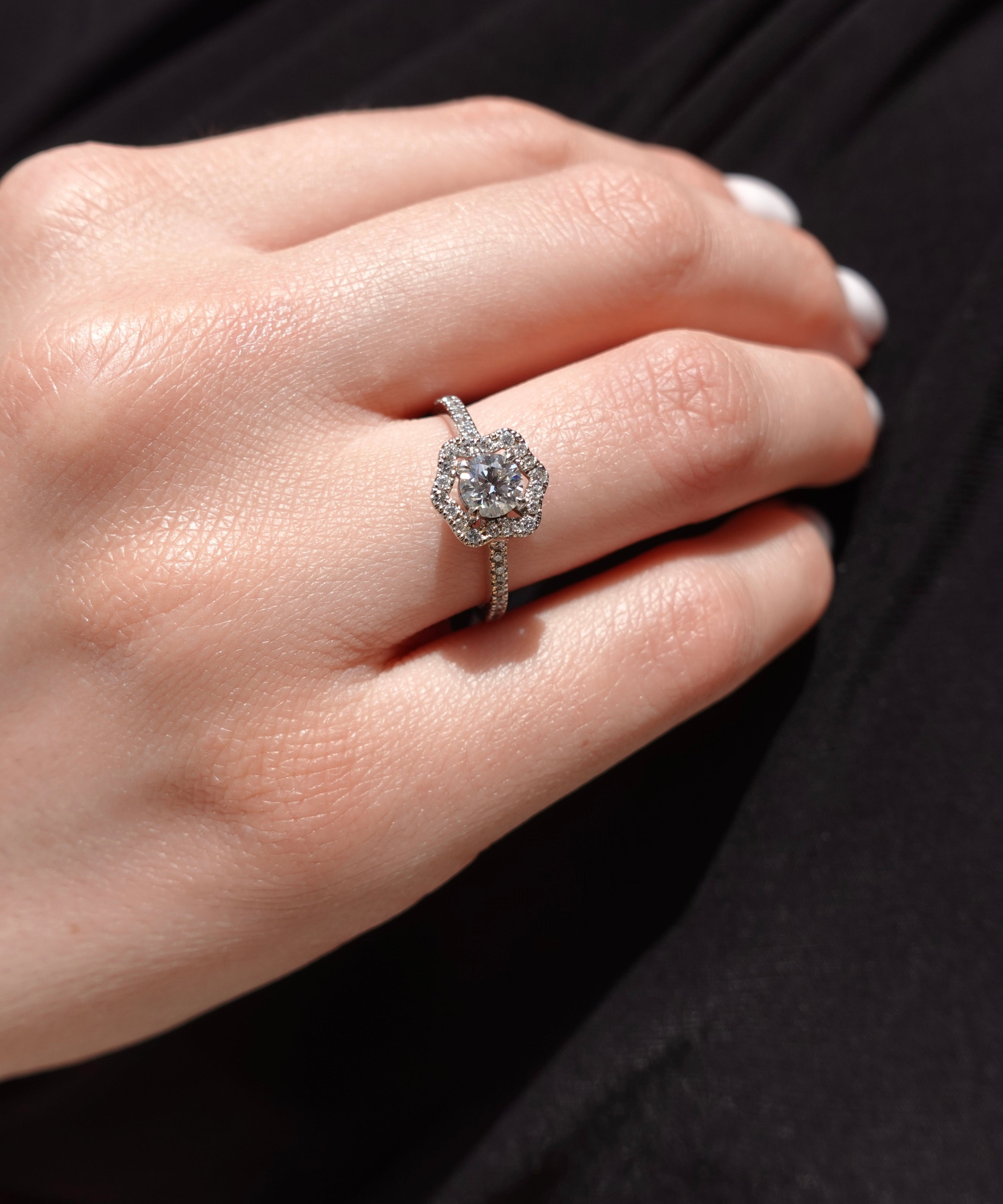 Close-up of a hand wearing an engagement ring with a diamond and flower-shaped halo on a dark background