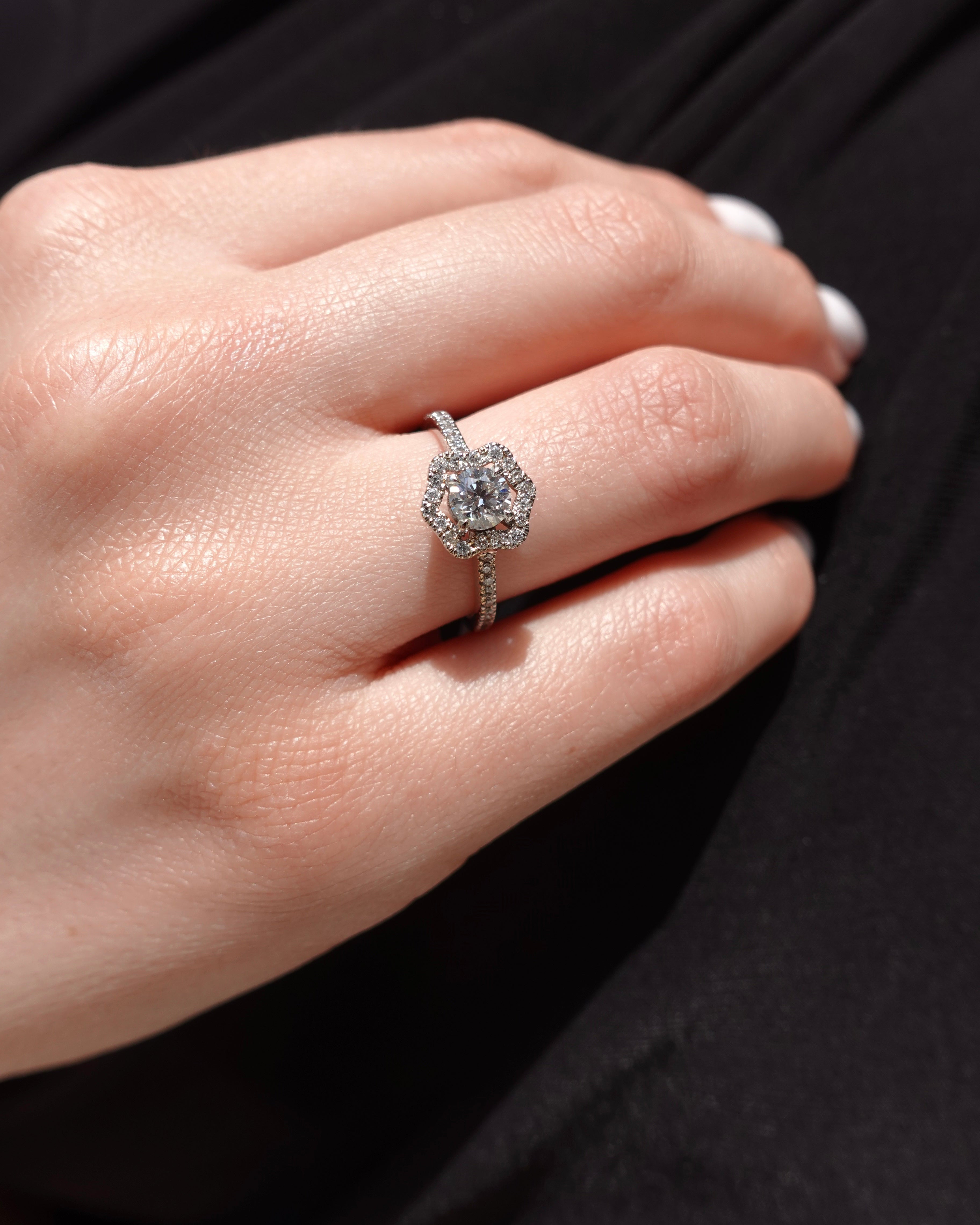 Close-up of a hand wearing an engagement ring with a diamond and flower-shaped halo on a dark background