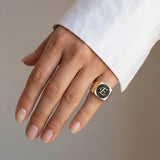 Hand wearing a gold ring with an initial E on a neutral background