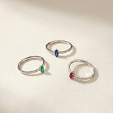 Three white gold rings with marquise-cut colored gemstones on a light beige background