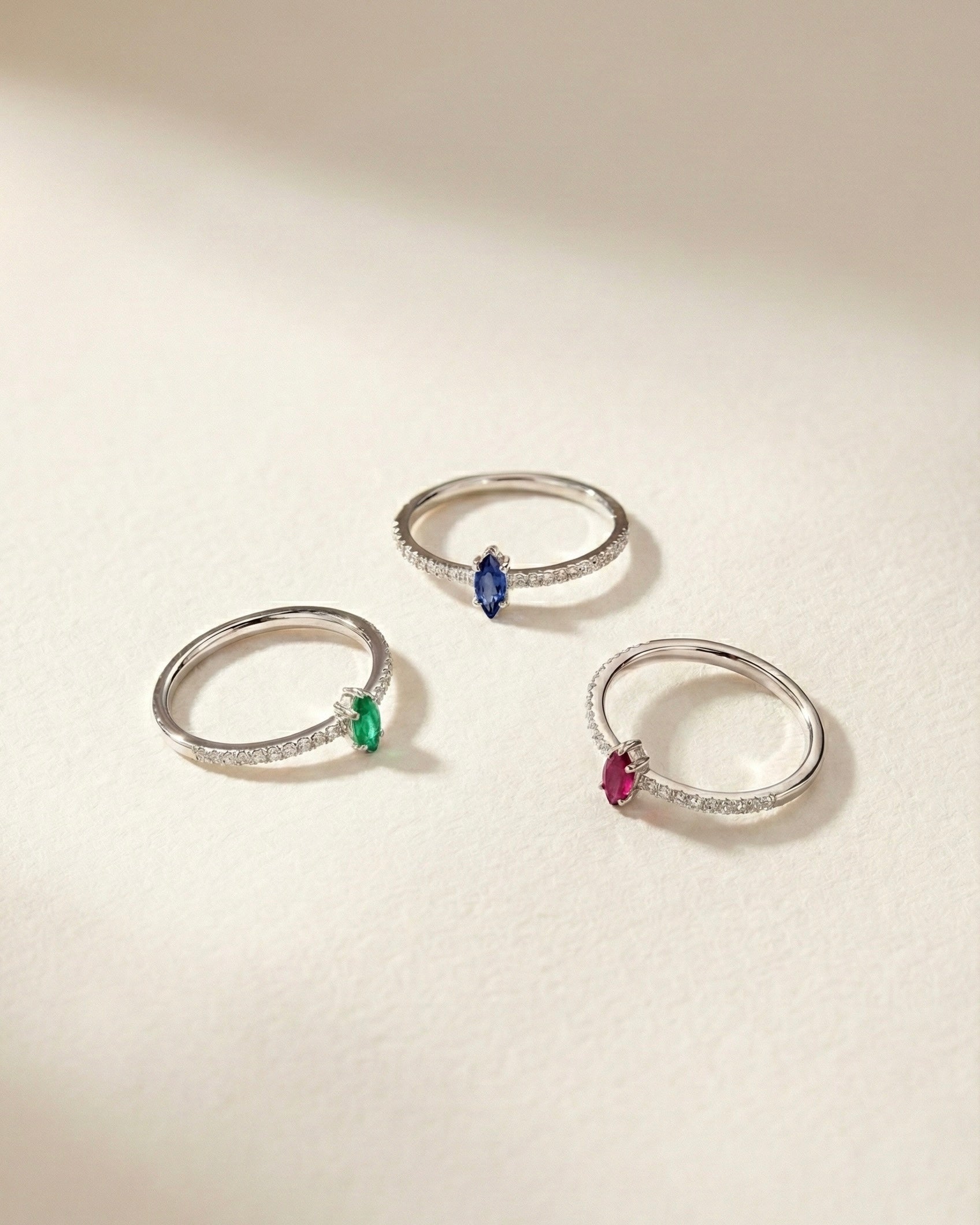 Three white gold rings with marquise-cut colored gemstones on a light beige background
