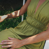 A woman wearing an olive green sleeveless dress with a deep V-neckline and a gathered smocked waist, seated outdoors. She wears a beautiful set of engagement ring and matching band. 