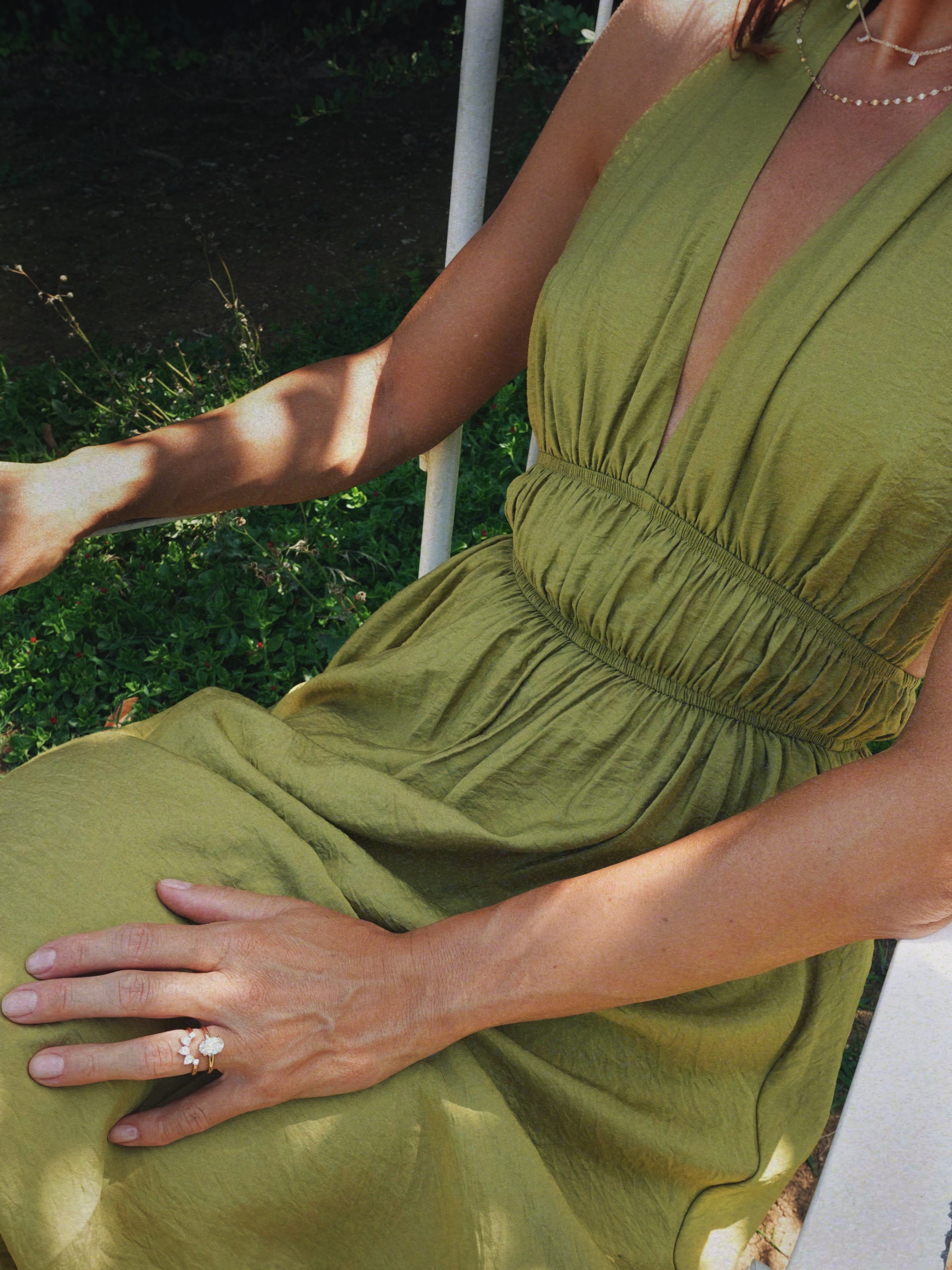 A woman wearing an olive green sleeveless dress with a deep V-neckline and a gathered smocked waist, seated outdoors. She wears a beautiful set of engagement ring and matching band. 