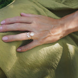 A close-up of a hand wearing a delicate gold engagement diamond ring with a white flower and leaf design, resting on draped olive green fabric.