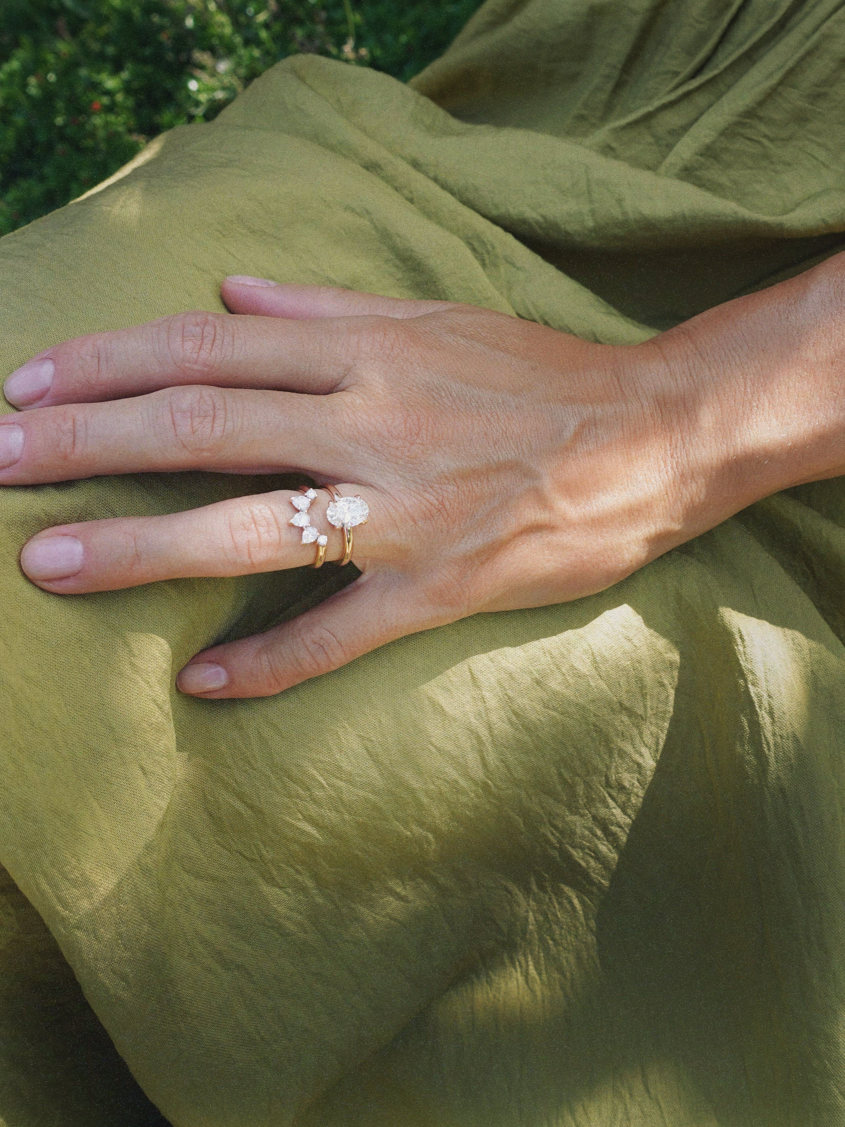 A close-up of a hand wearing a delicate gold engagement diamond ring with a white flower and leaf design, resting on draped olive green fabric.